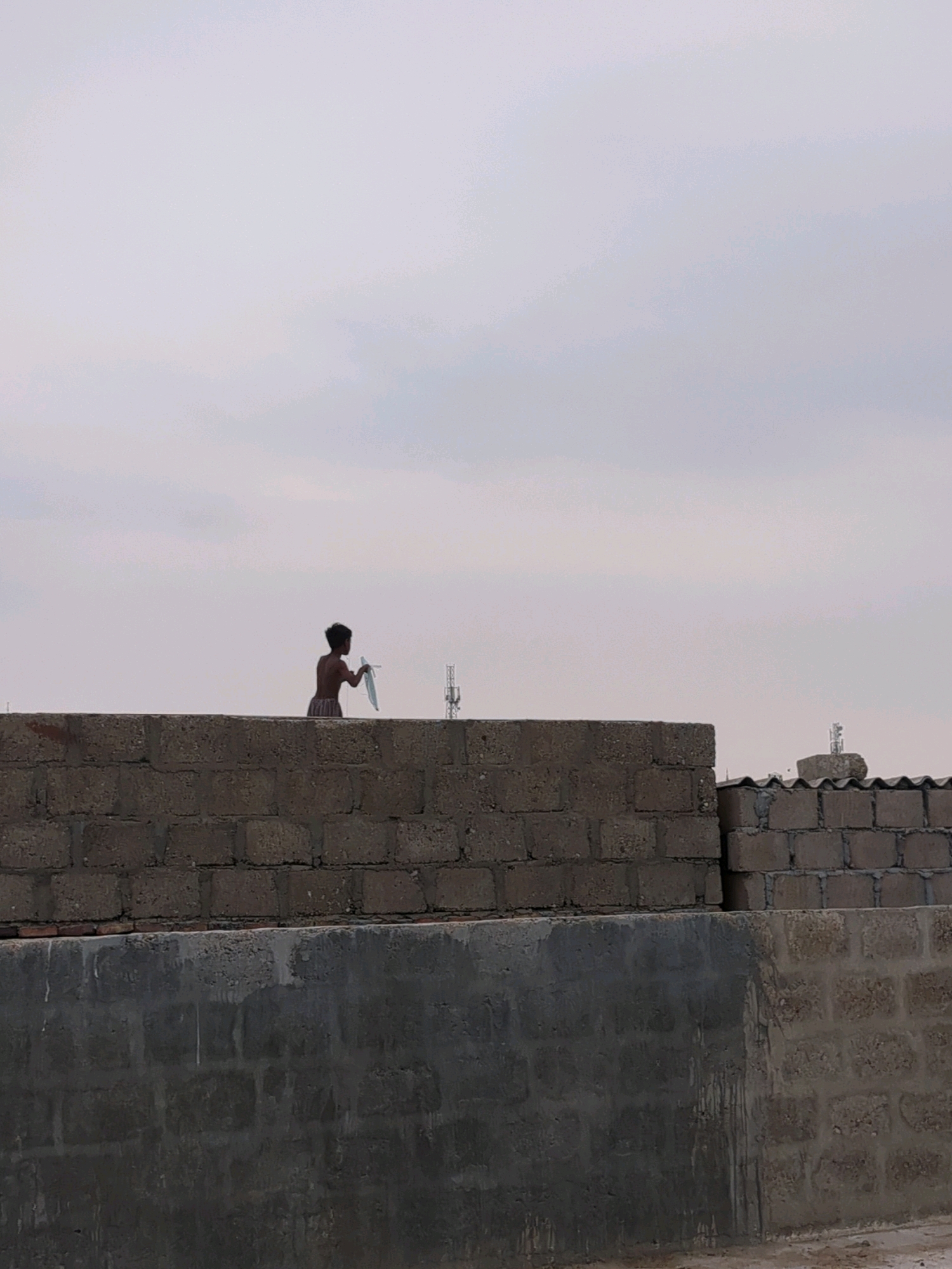 In Karachi, Pakistan, a young boy stands on a rooftop, holding a white kite. The boy, silhouetted against a cloudy sky, appears to be preparing to fly the kite. The rooftop is made of cinder blocks, and a concrete wall runs along the edge. In the background, a telecommunications tower is visible. The overall scene suggests a typical urban environment in Karachi, with a touch of childhood play.