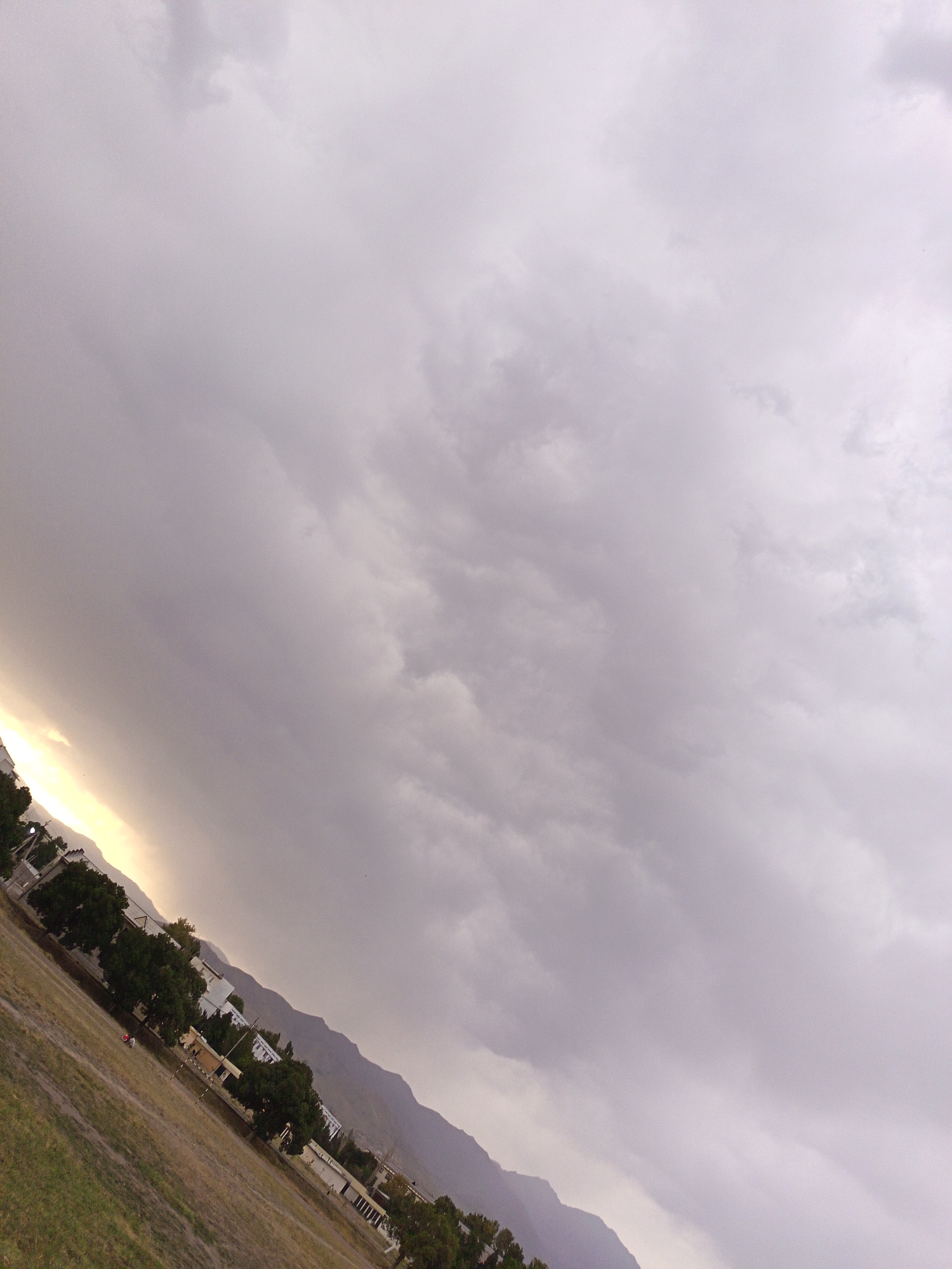 The image shows a wide landscape tilted to the right. The upper portion of the image is dominated by a cloudy sky with varying shades of grey and white. A bright, yellowish light is visible on the left side of the sky, suggesting a setting sun or a break in the clouds.
Below the sky, a mountainous landscape is depicted, with the peaks fading into the distance. A town or city is situated at the foot of the mountains, characterized by several buildings and dense green trees. The buildings appear to be low-rise structures. In the foreground, a large, open grassy field is visible, with some patches of dry, brown grass. Two small figures, possibly people, are present in the field, one in red clothing and another in white. The field appears to be a recreational area, perhaps a park or sports ground, with a long, thin structure that could be a goalpost or similar equipment. The overall impression is of a town or city located in a valley or mountainous region, under a dramatic sky.