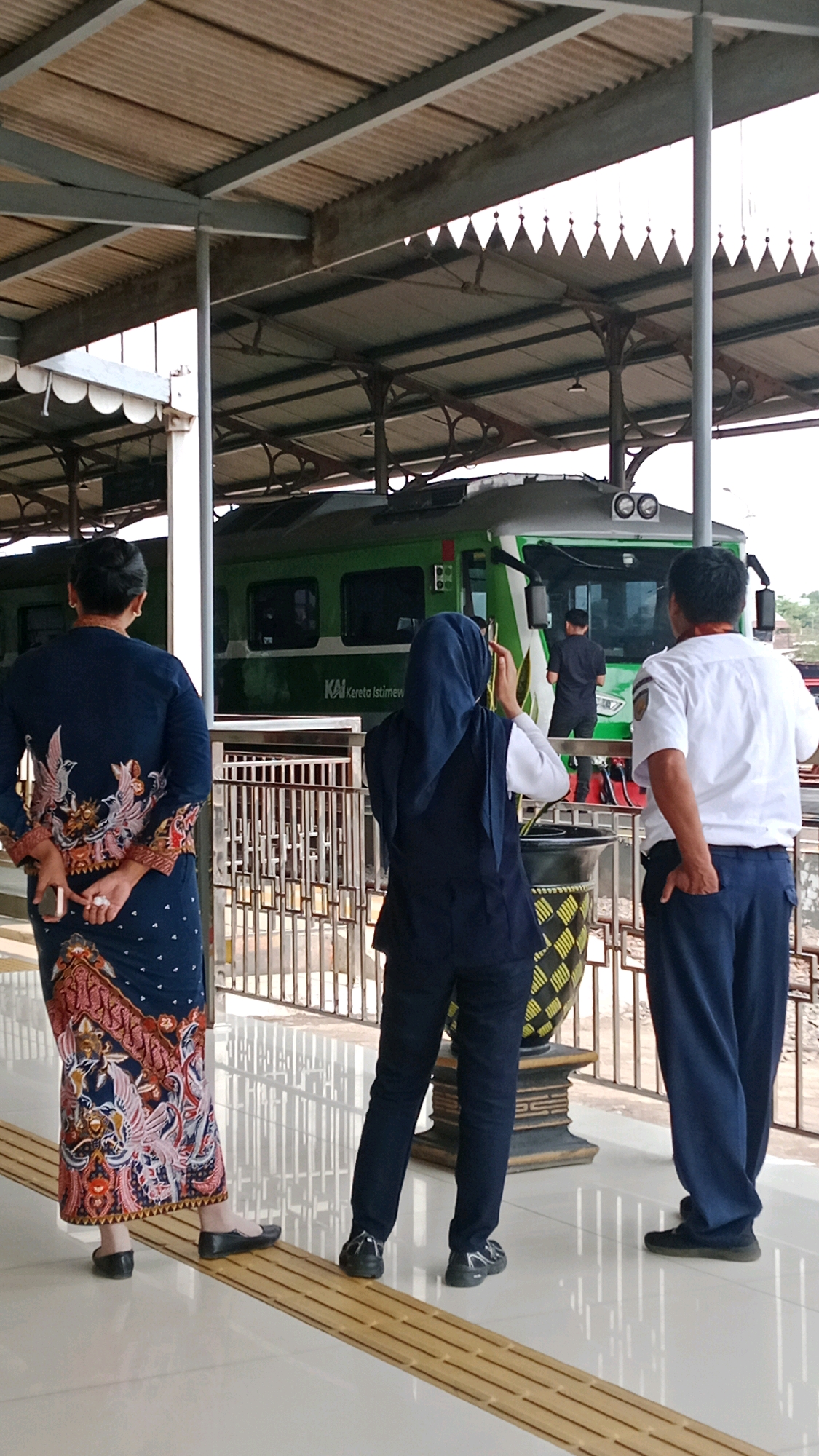 Three individuals stand on a train platform in Kidul Dalem, Indonesia, beneath a corrugated metal roof supported by metal beams and pillars. A green train, labeled "KAI Kereta Istimew," is visible in the background. The person on the left, viewed from behind, wears a dark blue, long-sleeved garment with ornate bird and floral patterns, and a dark blue skirt. They hold a smartphone in their left hand behind their back. In the center, another individual, also seen from behind, wears a dark blue hijab and a dark blue vest over a white shirt and dark pants, and is holding a smartphone to their face, possibly taking a photo. The person on the right, facing away from the camera, wears a white short-sleeved shirt and dark blue pants. They have their right hand in their pants pocket. The platform has a tactile warning strip made of yellow tiles running horizontally across the foreground. A decorative planter with a patterned base is situated between the two individuals in the center and right. The lighting suggests it is daytime.