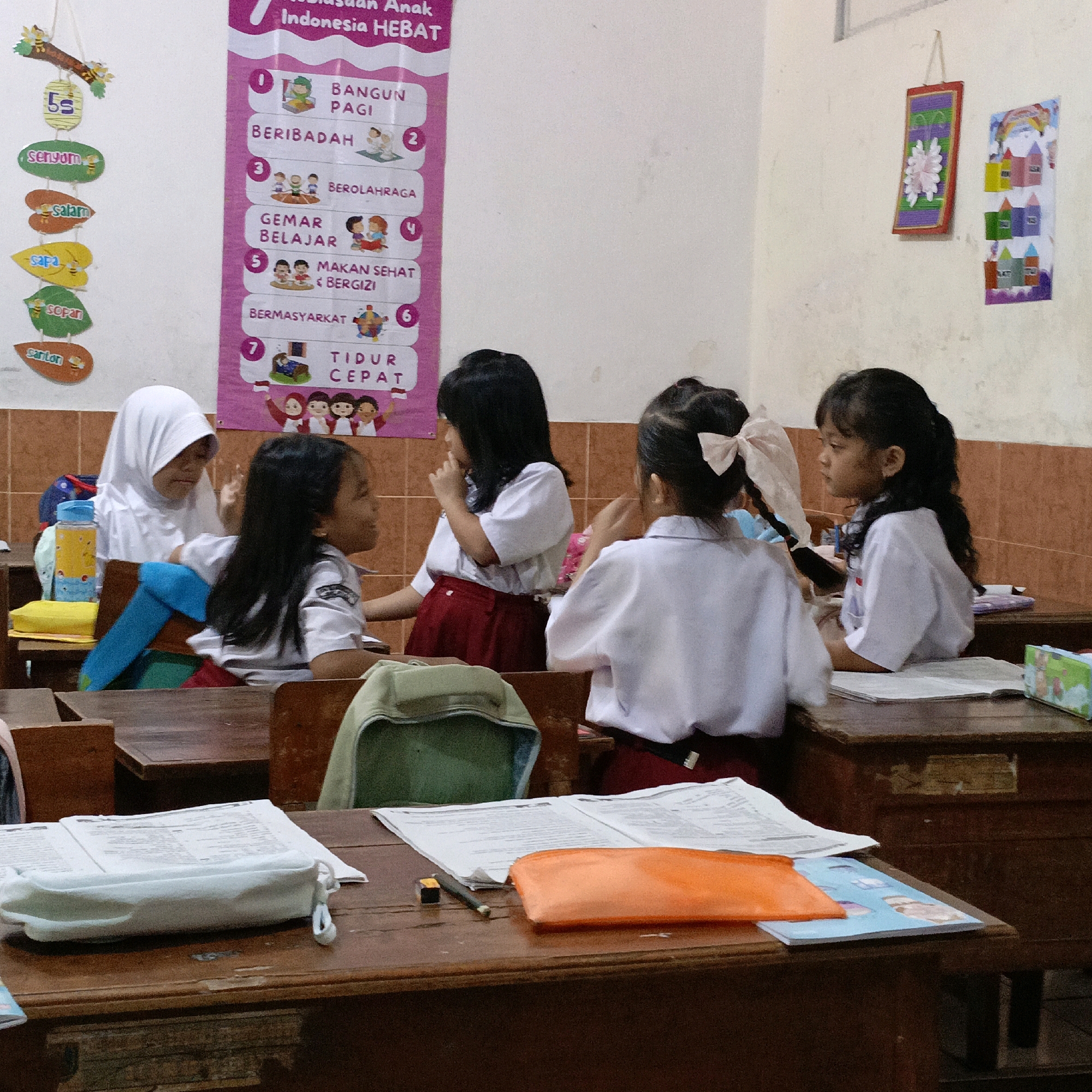 In a classroom in Salatiga, Indonesia, four young schoolgirls in white uniforms and red skirts are seated at wooden desks. One girl, wearing a white hijab, is looking down. Another girl to her left is looking to her right, her mouth open as if speaking. A third girl, facing away from the camera, has her hand near her mouth and appears to be listening. The fourth girl on the right is also looking to her right, her head tilted. On the wall behind the students, colorful posters are displayed, one with a list of rules or values for "Anak Indonesia HEBAT" (Great Indonesian Children) and another with drawings of houses and a rainbow. The desks are cluttered with school supplies, including open notebooks, pencil cases, and a water bottle. The room has tiled walls and wooden furniture, suggesting a typical Indonesian elementary school setting.