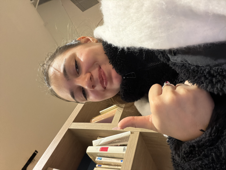 A woman in a black fleece jacket and white scarf smiles, positioned to the left of a light-colored wooden bookshelf with books and a pink object on the shelves. She gives a thumbs-up, displaying a ring with a red stone on her right hand. The scene is lit with warm lighting.