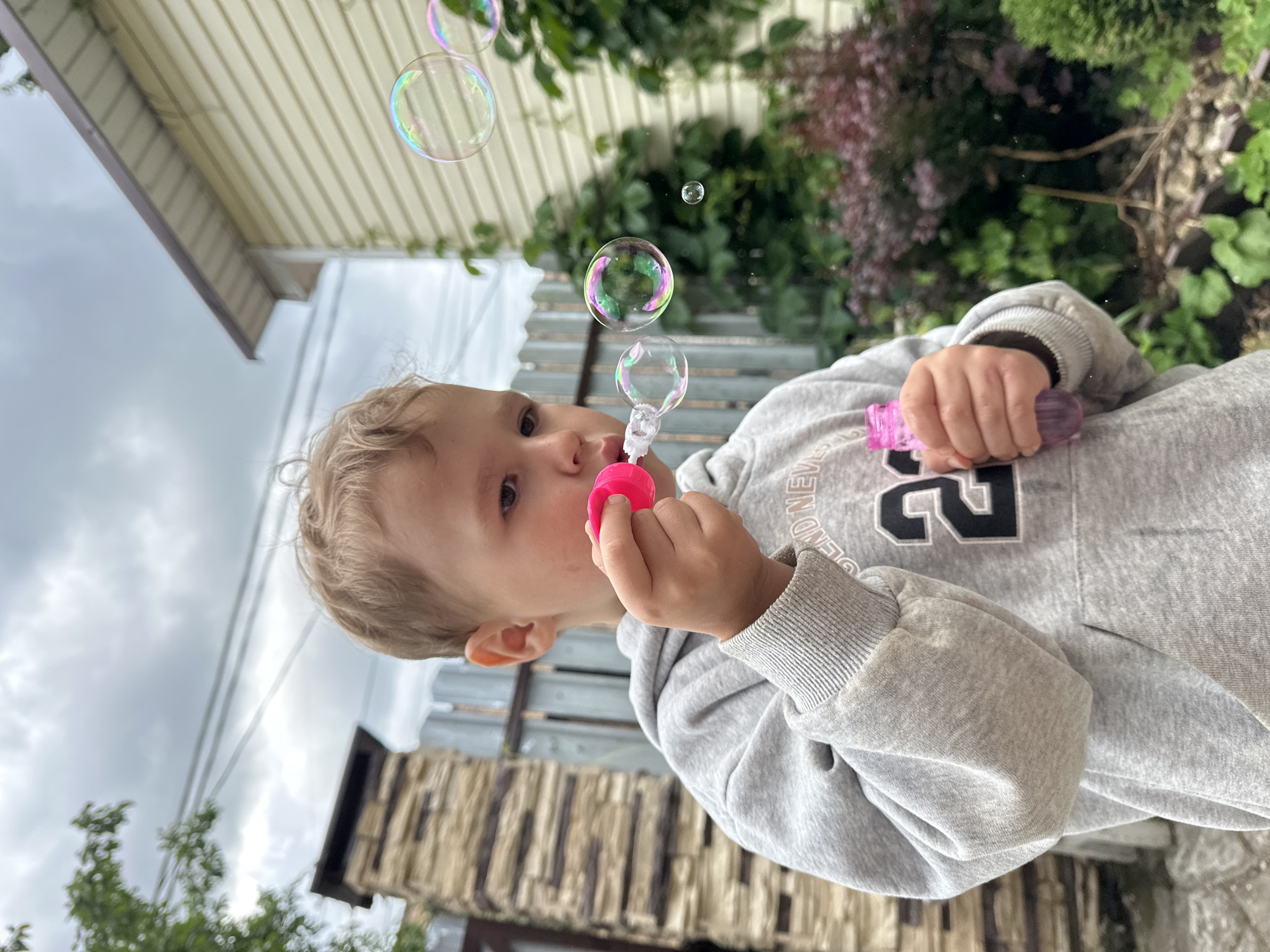 A young boy is blowing bubbles in a backyard in Nizhny Novgorod, Russia. He is wearing a gray hooded sweatshirt with a black number "24" on it. He holds a pink bubble wand in his right hand and a pink bottle of bubble solution in his left. Several iridescent bubbles are floating in the air. In the background, there is a light-colored house with a textured wall, a stone pillar, a wooden fence, and green bushes. The sky is cloudy.