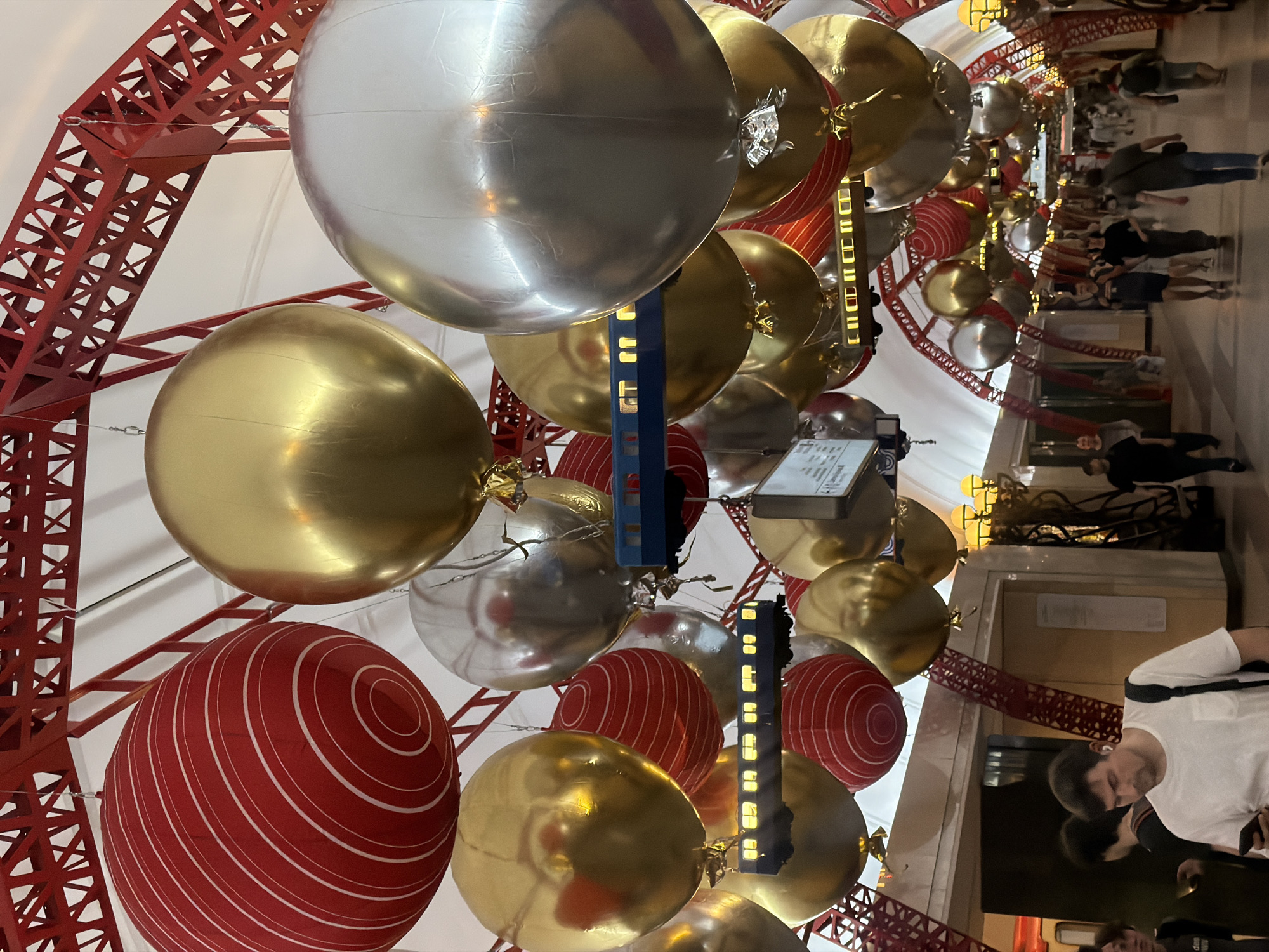 The image shows an indoor scene with a low angle view looking up at a decorated ceiling and a blurred crowd of people in the background. The ceiling structure appears to be a red, metal framework resembling the Eiffel Tower. Hanging from the framework are large, spherical ornaments in gold, silver, and red with white spiral patterns. There are also dark, rectangular objects with small, illuminated squares attached to the structure. In the background, a crowd of people is walking in a blurred, horizontal line. The lighting is bright and diffused, suggesting an interior space with ample artificial illumination.
The location is inside the GUM department store in Moscow, Russia. The decorations are holiday themed.
