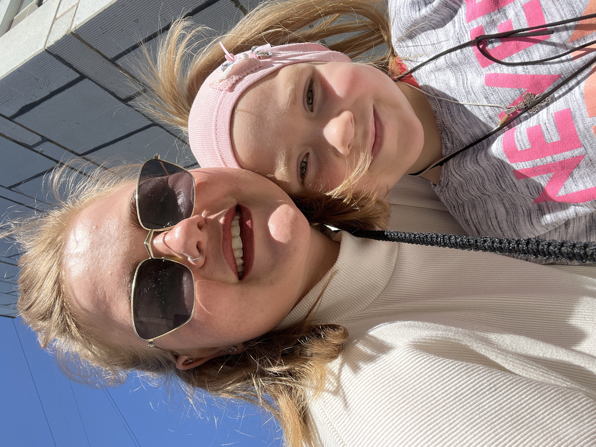A selfie taken from a low angle shows a woman and a young girl. The woman is wearing sunglasses and a white shirt. The girl has a pink headband and a gray shirt with pink letters. The sky and a building are visible in the background. There is no specific information in the image that identifies the location as Chaikovsky, Russia.