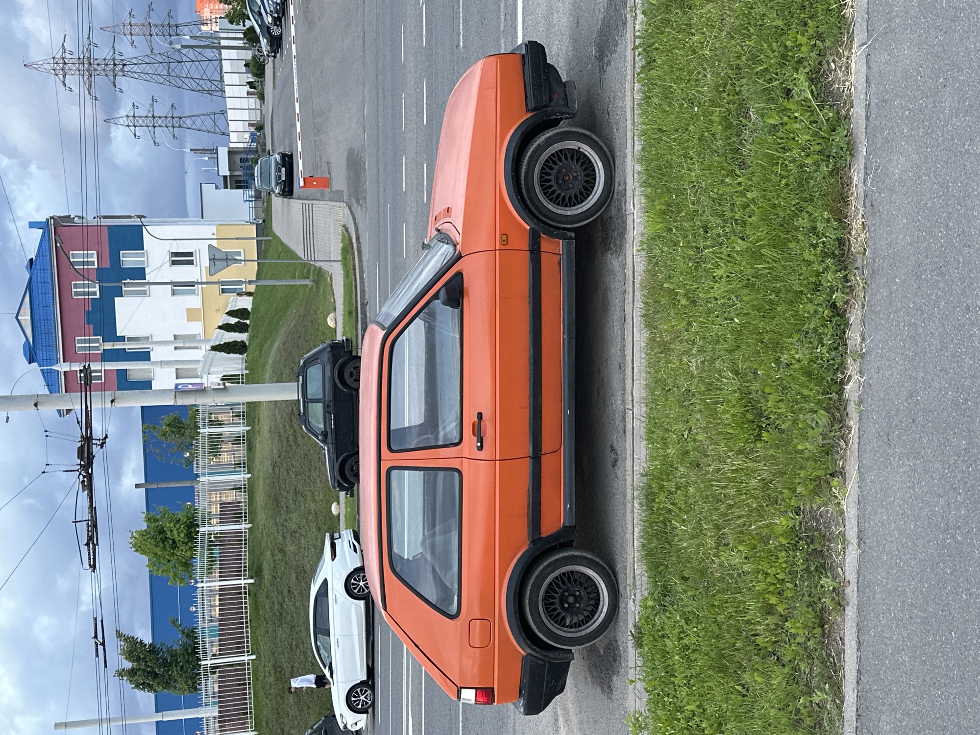 The image depicts a bright orange Volkswagen Golf MK2 parked on the side of a paved road, partially on the grass verge. Alongside it are other parked vehicles, including a white hatchback and a black SUV. In the background, there is a multi-colored building with a blue roof and a white fence running parallel to the road. Overhead power lines are visible against a cloudy sky. The location is identified as Minsk, Belarus based on recognizable landmarks and the specific arrangement of elements in the scene.
