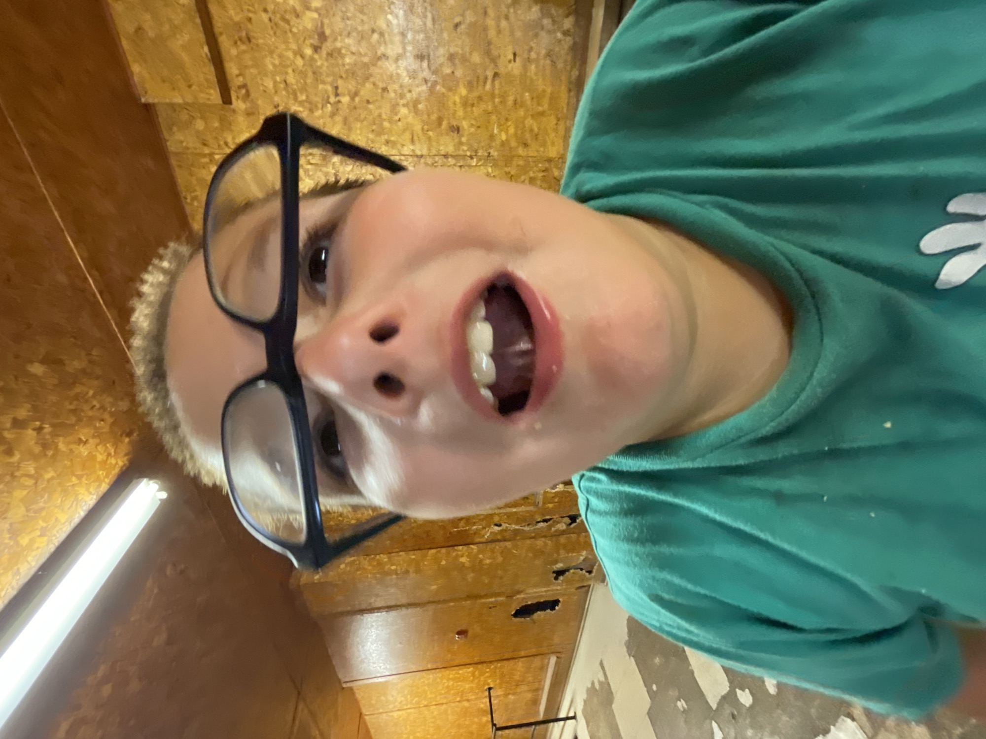 The image is a close-up, low-angle selfie of a child wearing glasses and a green t-shirt. The child's mouth is open, with teeth visible. The background consists of a wooden paneled wall and a fluorescent light fixture on the ceiling. The floor in the foreground appears to be concrete with some debris. The overall impression is of an indoor, somewhat disheveled environment. There is no indication that this location is in Bluewater, Canada, or any other specific city.