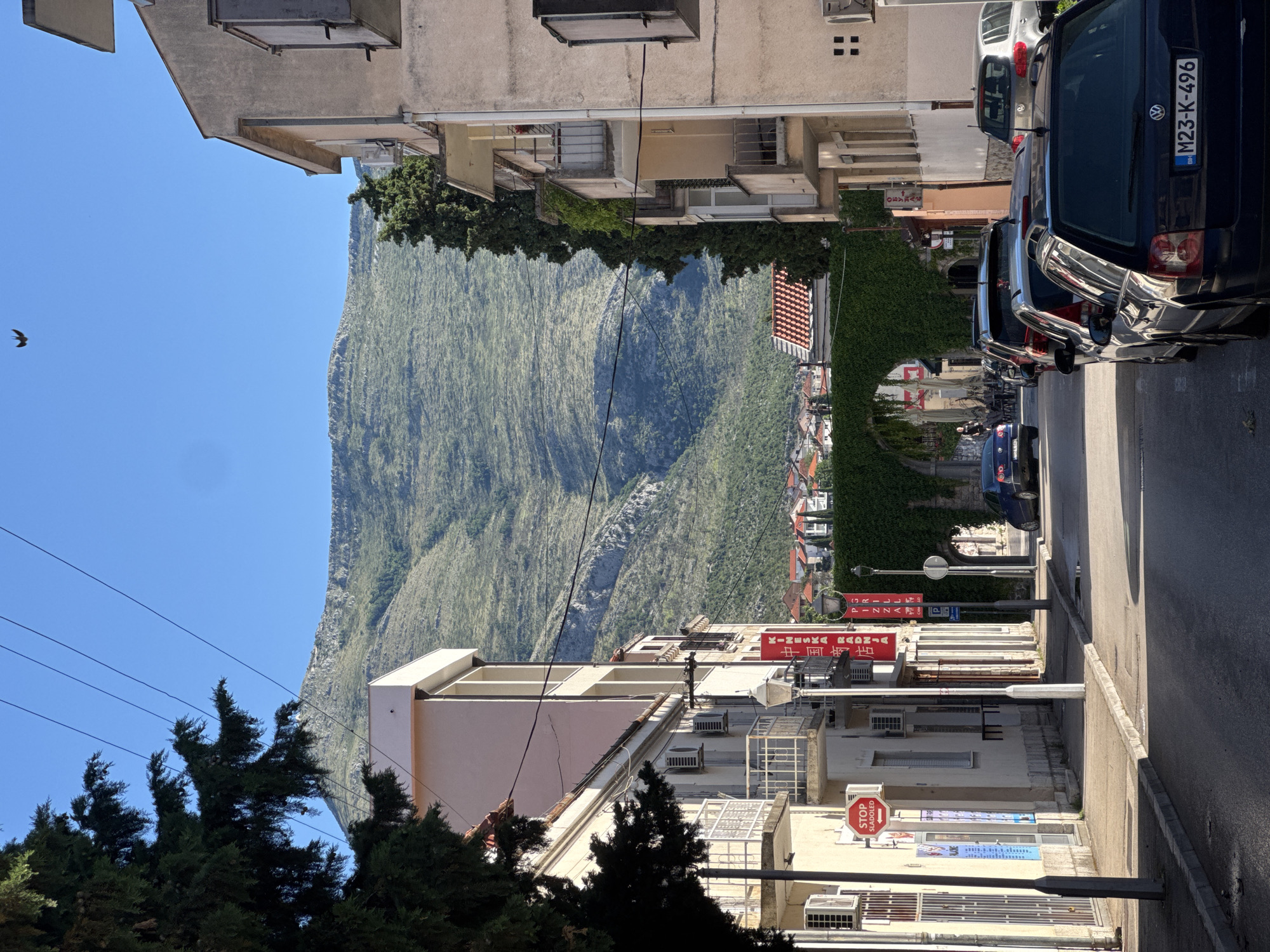 The image shows a street in Trebinje, Bosnia and Herzegovina, with buildings on both sides and a mountain in the background. On the left, there is a light-colored building with balconies and air conditioning units. A red sign with white text reading "STOP SLADOLED" (Stop Ice Cream) is visible. Further down the street, a green, arched structure covered in ivy is seen, with a few cars parked or driving past it. The right side of the street features a similar building with balconies and architectural details. A dark-colored car with a license plate reading "M23-K-496" is parked in the foreground. The sky is clear and blue, and the sun casts shadows on the street and buildings, indicating it is daytime. The overall scene depicts a residential and commercial street in a Mediterranean-like environment.