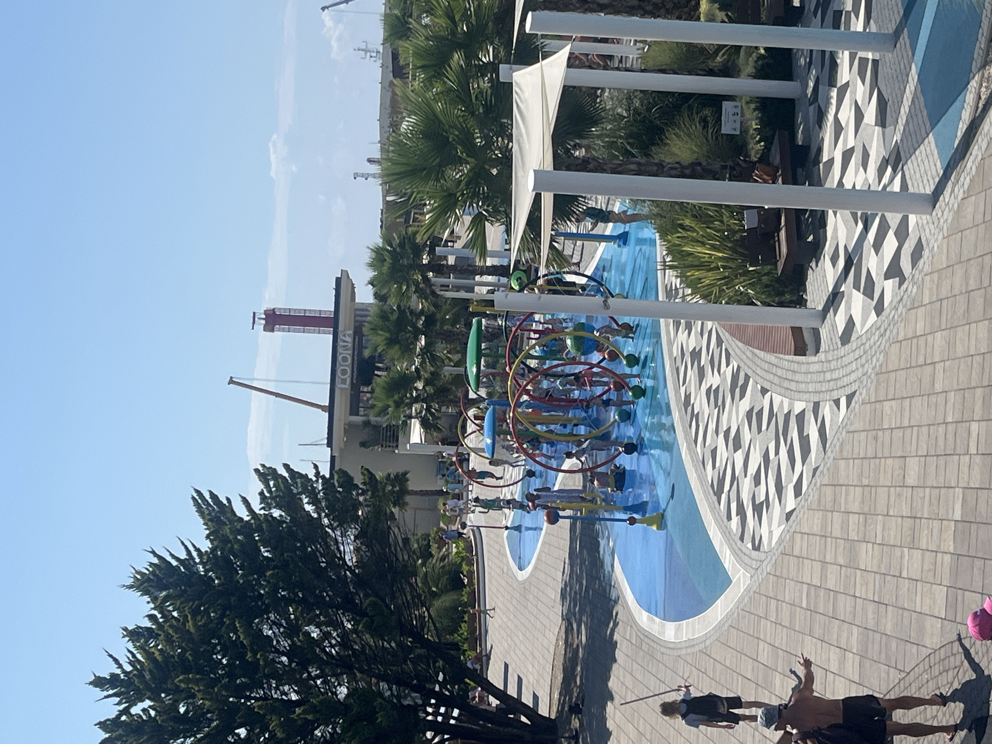 The image depicts a water park in Sochi, Russia, likely during daytime and under clear skies. In the foreground, a person is seen walking away from the camera, and another person is seen from behind, facing away and gesturing with their right arm. Further in the mid-ground, a children's water play area is active with several children engaged in water activities. This area features colorful, circular spray structures with various water nozzles. Surrounding the water park is a paved pathway with a geometric pattern, and lush green vegetation, including palm trees. In the background, a building with the word "LOONA" is visible, and a tall, red structure, possibly a tower, stands against the sky. The overall scene suggests a recreational area on a sunny day.