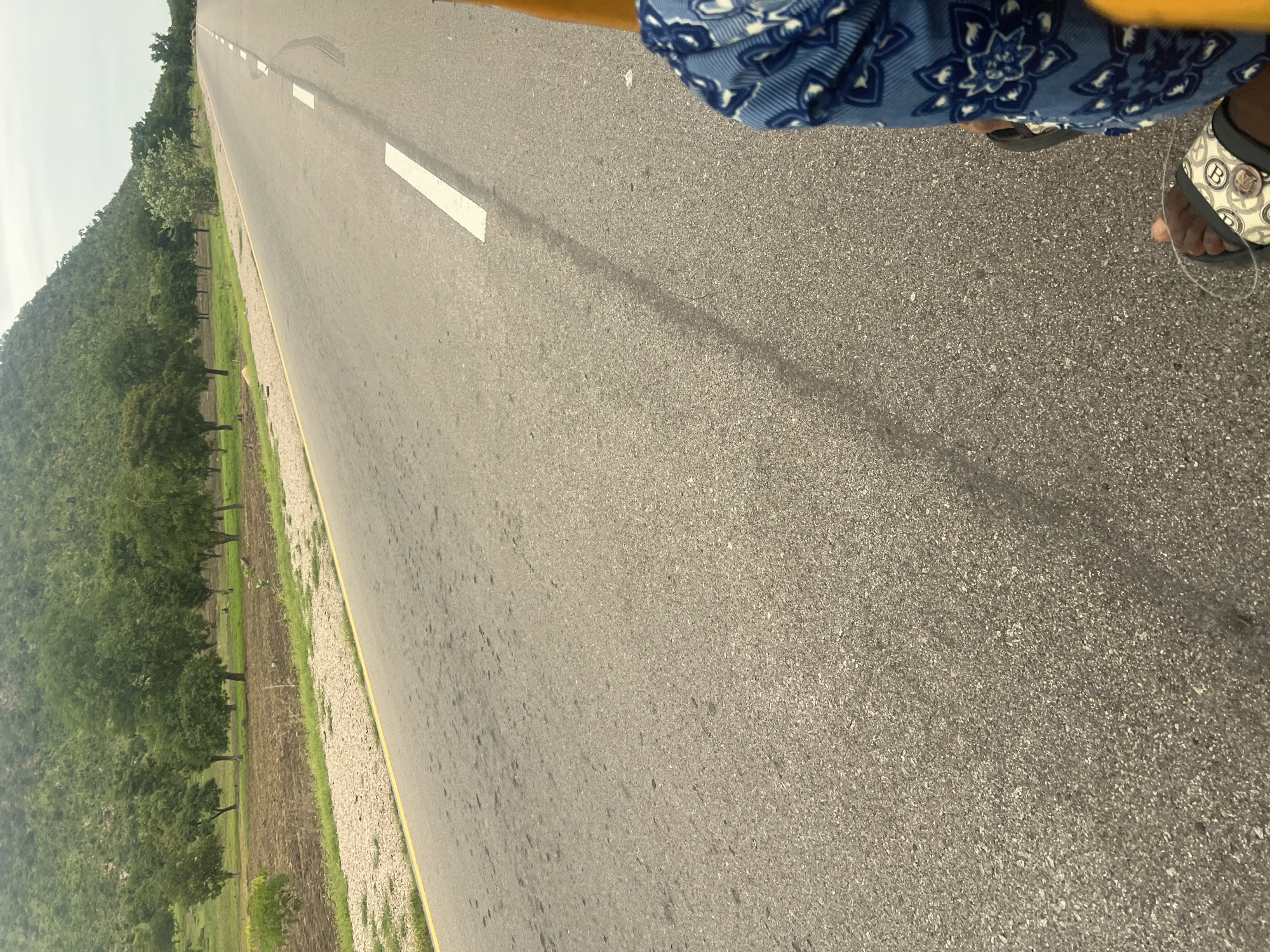 The media file shows a road scene from a tilted, ground-level perspective, suggesting the camera is being held or is positioned low. The road, paved with asphalt, dominates the right side of the frame, stretching into the distance with a visible white lane marking. To the left of the road, a grassy shoulder transitions into a field and then a densely forested hillside, suggesting a rural or natural setting. The sky is overcast, indicating a cloudy day, and the lighting suggests it's daytime, likely mid-morning or afternoon.
In the foreground on the right, a person's feet are visible, wearing patterned blue fabric on their upper legs and a pair of sandals. One sandal is a dark color, while the other is white with a distinctive pattern featuring the letters "LV" arranged in a circular design. The person appears to be standing or walking on the roadside, with their left foot closer to the camera. The overall mood is mundane, capturing a moment of travel or rest in a scenic, possibly remote, location. There is no visible text in the image, beyond the branding on the sandal. The location context provided is Minda, Nigeria.