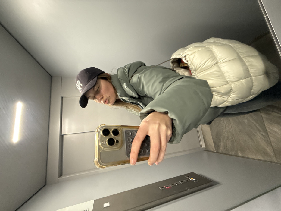 The image displays a selfie taken by a young person inside what appears to be an elevator. The photograph is oriented sideways, making the subject and the elevator walls appear horizontal.
The main subject is a person wearing a dark baseball cap with a prominent white "NY" (New York Yankees) logo on the front. They have long, light-colored hair peeking out from under the cap. They are dressed in a thick, sage green puffer jacket. Their eyes are closed or squinting, and their lips are playfully pursed in a "duck face" expression. The person is holding a gold or champagne-colored smartphone in their right hand, clearly visible in the foreground. The phone has a distinct multi-lens camera module and is protected by a transparent, somewhat bulky case with a textured, ribbed grip on its right side.
Behind the person, a large, cream-colored or off-white puffer jacket is visible, suggesting either it's being carried or another person is partially present but obscured. The background consists of smooth, light gray walls, typical of an elevator interior. On the left side of the image, a vertical strip of bright white light illuminates the wall. At the bottom of the frame (which would be the floor if correctly oriented), an elevator control panel is visible. This dark gray panel features multiple light-colored buttons, with some displaying illuminated red or orange indicators. The floor surface appears to be a darker gray or mottled pattern. The overall lighting suggests an indoor, artificial light source.