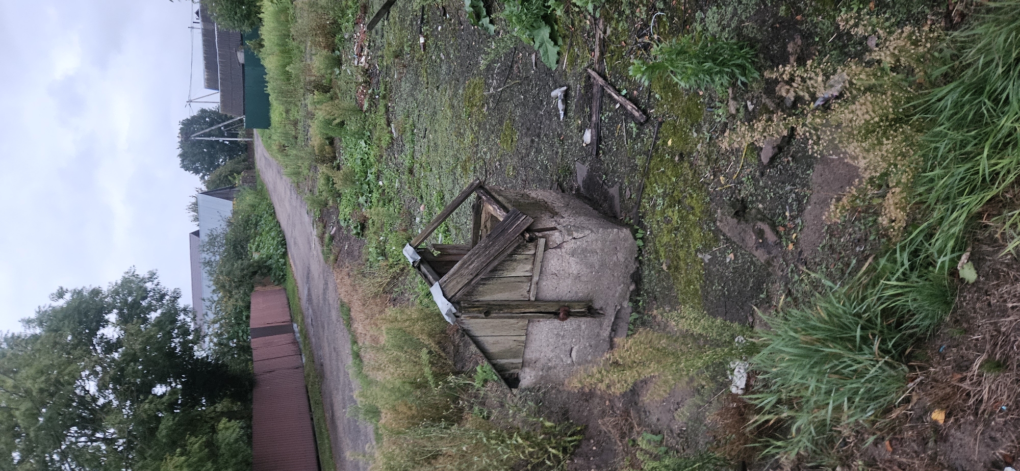 The image depicts an outdoor scene in a rural or suburban setting, likely in Liozna, Belarus, under an overcast sky. In the foreground, a weathered, square concrete wellhead with a dilapidated wooden roof structure is visible. The well is surrounded by overgrown grass and weeds. To the left, a dark brown corrugated metal fence runs along a dirt path or road. Beyond the fence, a line of trees and foliage is present. The road curves into the distance, flanked by more vegetation and the roofs of several buildings, some with metal roofing. Some debris is scattered on the ground near the wellhead and along the road. The overall impression is one of a neglected or abandoned area.