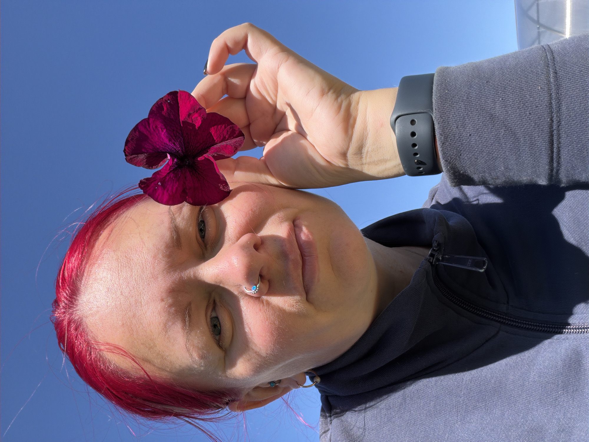 A woman with bright pink hair looks into the camera, holding a dark purple flower in her right hand. She is wearing a grey zip-up jacket, a nose ring with a blue gem, and earrings. Her left hand is resting on the lapel of her jacket. The background is a clear blue sky. The image was taken outdoors in Chaikovsky, Russia.