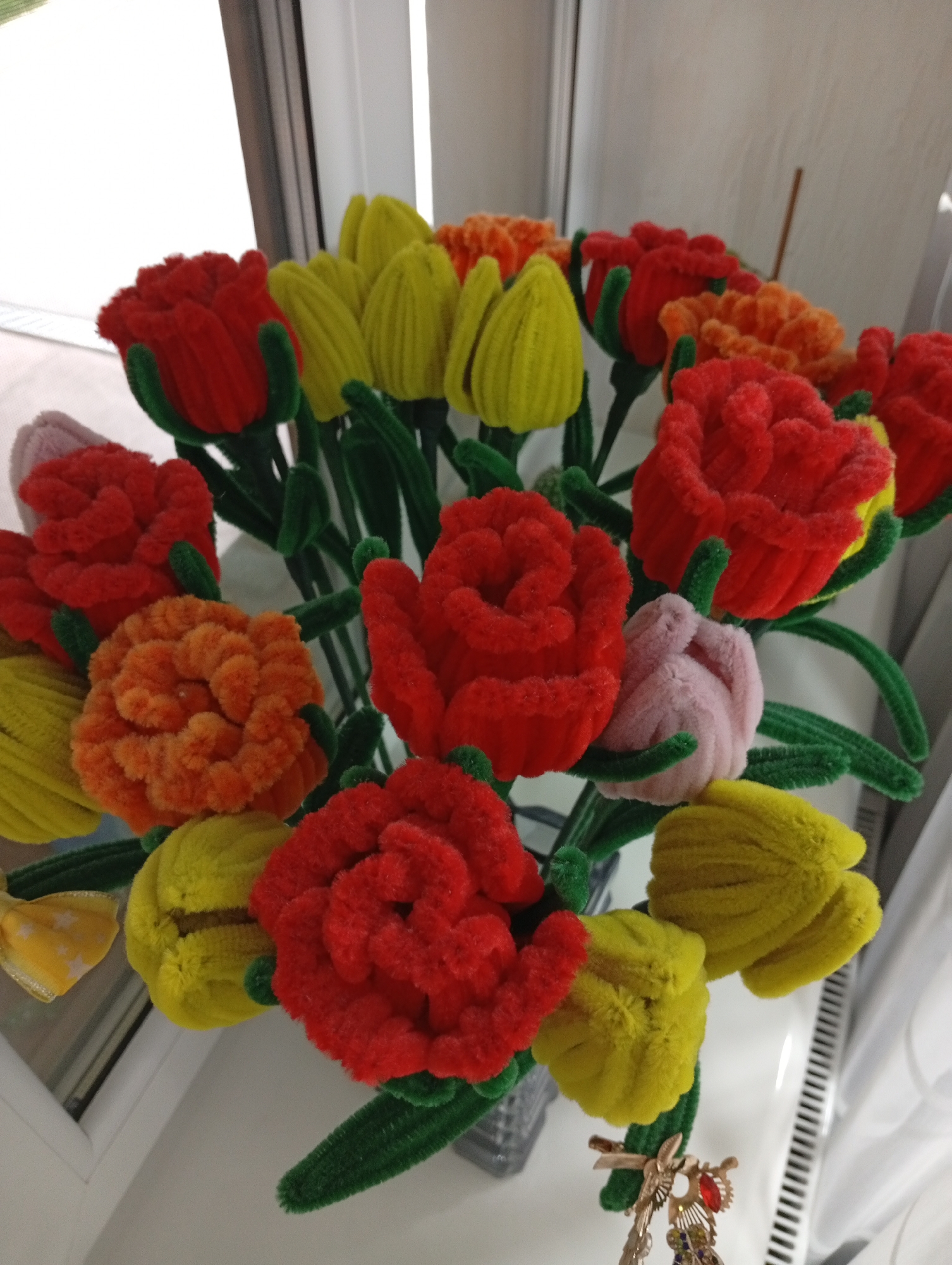 The image shows a bouquet of artificial flowers, specifically tulips and roses, made from fluffy material. The flowers are in various colors, including red, yellow, orange, and pink. They are arranged in a vase, which is partially visible at the bottom center of the image. The bouquet is placed on a white surface, possibly a windowsill, next to a window with vertical blinds. The overall lighting suggests an indoor setting. There is a decorative charm hanging from one of the flower stems. There is no discernible location within the city of Pănășești, Moldova indicated in the image.