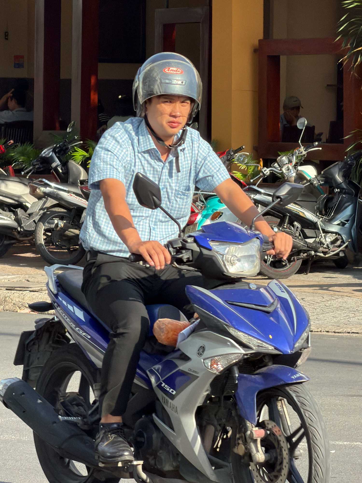 A young Vietnamese man wearing a blue and white checkered shirt, black pants, and a silver helmet is riding a blue and silver motorcycle in Nha Trang, Vietnam. He is looking towards the camera. The motorcycle has the brand name "YAMAHA" visible on its side. In the background, there are other motorcycles parked and a building with vertical brown and white stripes and a brown frame around a window. Sunlight is casting shadows on the ground and the subjects. The scene appears to be on a street in Nha Trang.