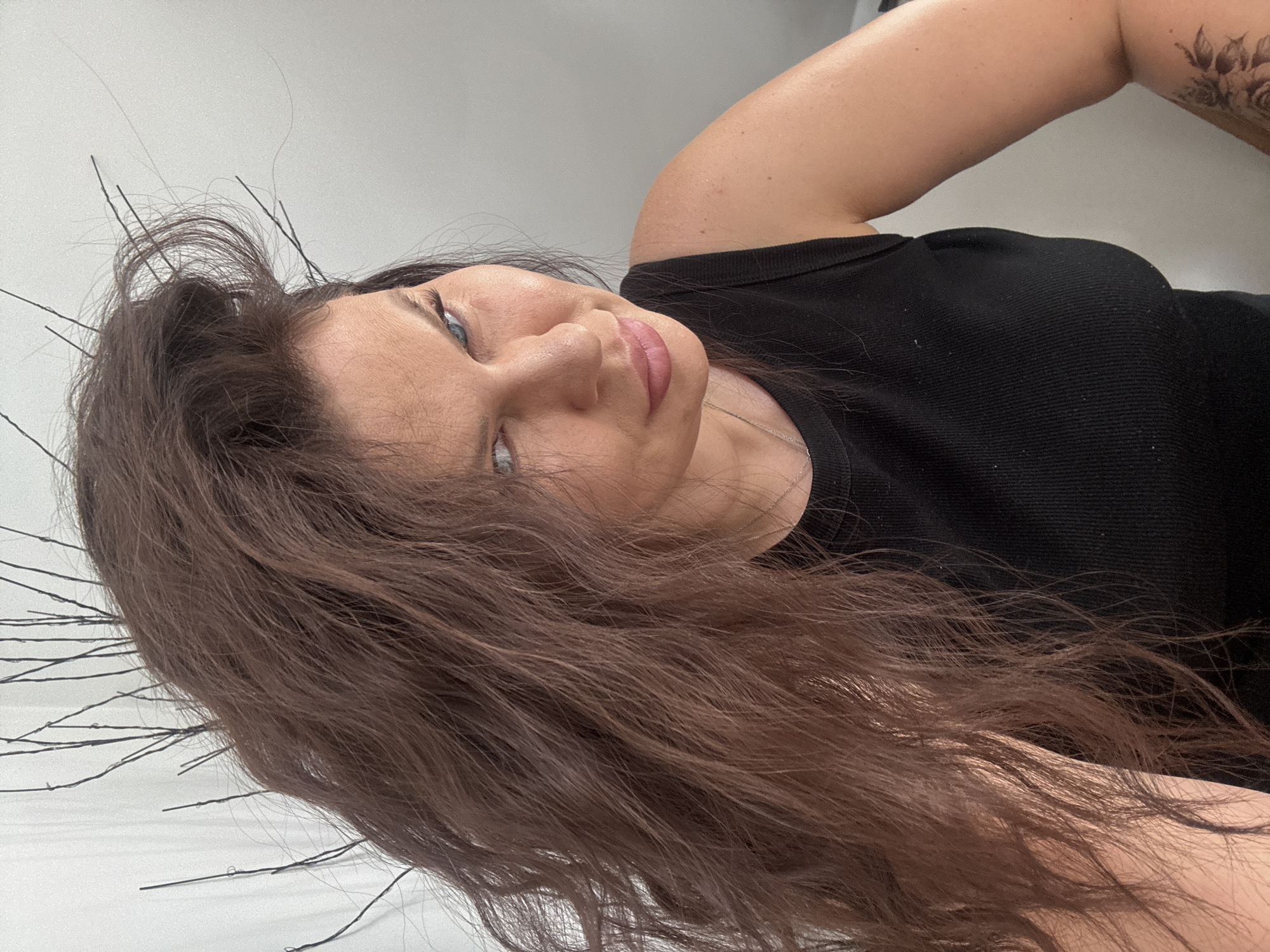 A woman with long, brown, wavy hair is looking at the camera with a slight smirk. She is wearing a black tank top and a silver necklace. Her hair is partially obscuring her face, with some thin, dark branches sticking out from behind it. The background is a plain, light-colored wall, possibly white or off-white. The lighting suggests an indoor setting. The woman's left shoulder and arm are visible, and a floral tattoo can be seen on her upper arm. Her eye color appears to be blue. The image was taken in Katowice, Poland.