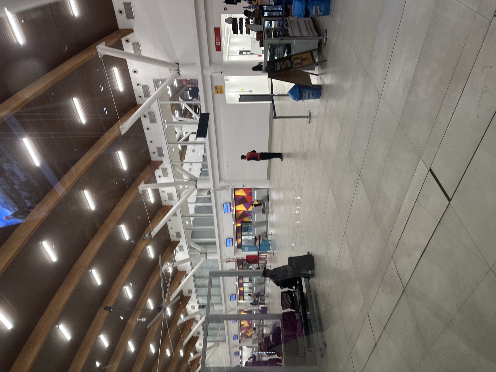 The image depicts the interior of an airport in Arica, Chile. The space is well-lit with a high, arched wooden ceiling featuring rows of fluorescent lights. The floor is tiled in a grid pattern with light grey tiles. The walls are predominantly white with some sections painted in bright, geometric patterns.
A person wearing a black uniform with a red vest stands near a wall. Further away, there are check-in counters. In the foreground, there is a luggage cart with luggage, a seated individual and a glass barrier.