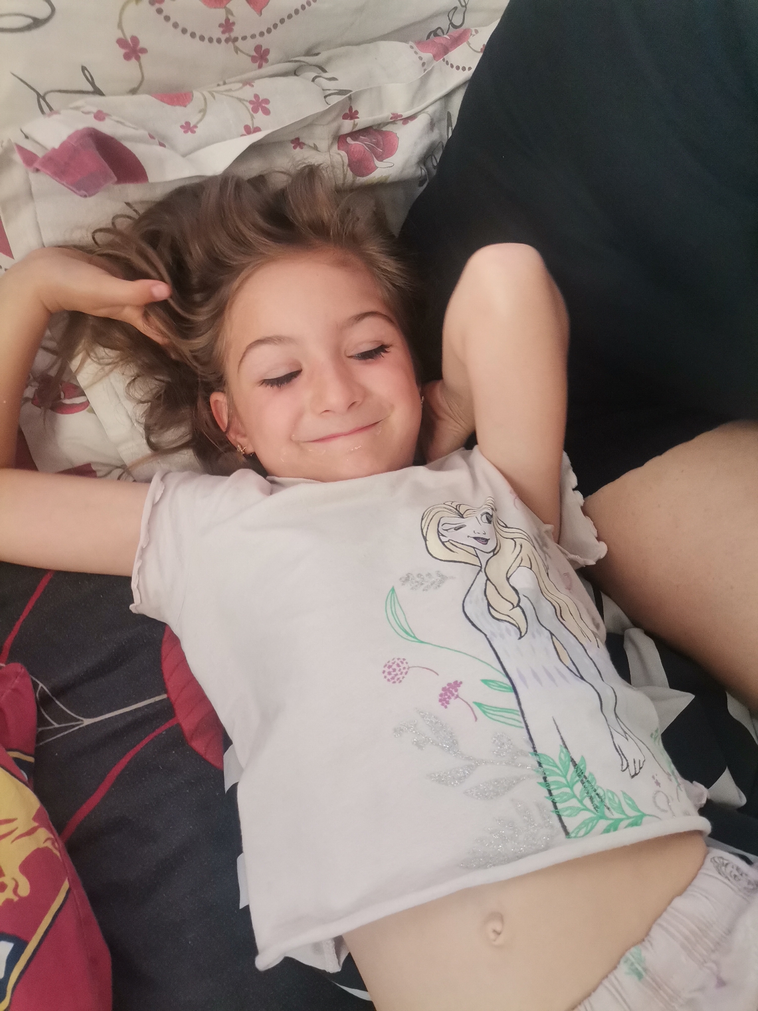 A young girl with long, wavy brown hair lies supine, her arms bent with hands clasped behind her head, in a relaxed pose. She is smiling broadly with her eyes closed, suggesting happiness or contentment. A small amount of liquid or residue is visible around her mouth. She wears a light-colored t-shirt featuring an illustration of Elsa from Disney's Frozen, characterized by long blonde hair, a light blue/purple dress design, and green foliage with glitter accents. Below her t-shirt, light-colored, striped patterned shorts or underwear are visible, and her navel is exposed.
She is situated on a bed or similar surface, with her head resting on a white pillow adorned with pink floral patterns. To the top right, a dark, solid-colored fabric covers what appears to be part of an adult's leg or torso, suggesting another person is present or nearby. The lower part of the image shows a darker patterned blanket or sheet with red, black, and white designs. The setting appears to be indoors, likely a bedroom. There is no visible text in the image.