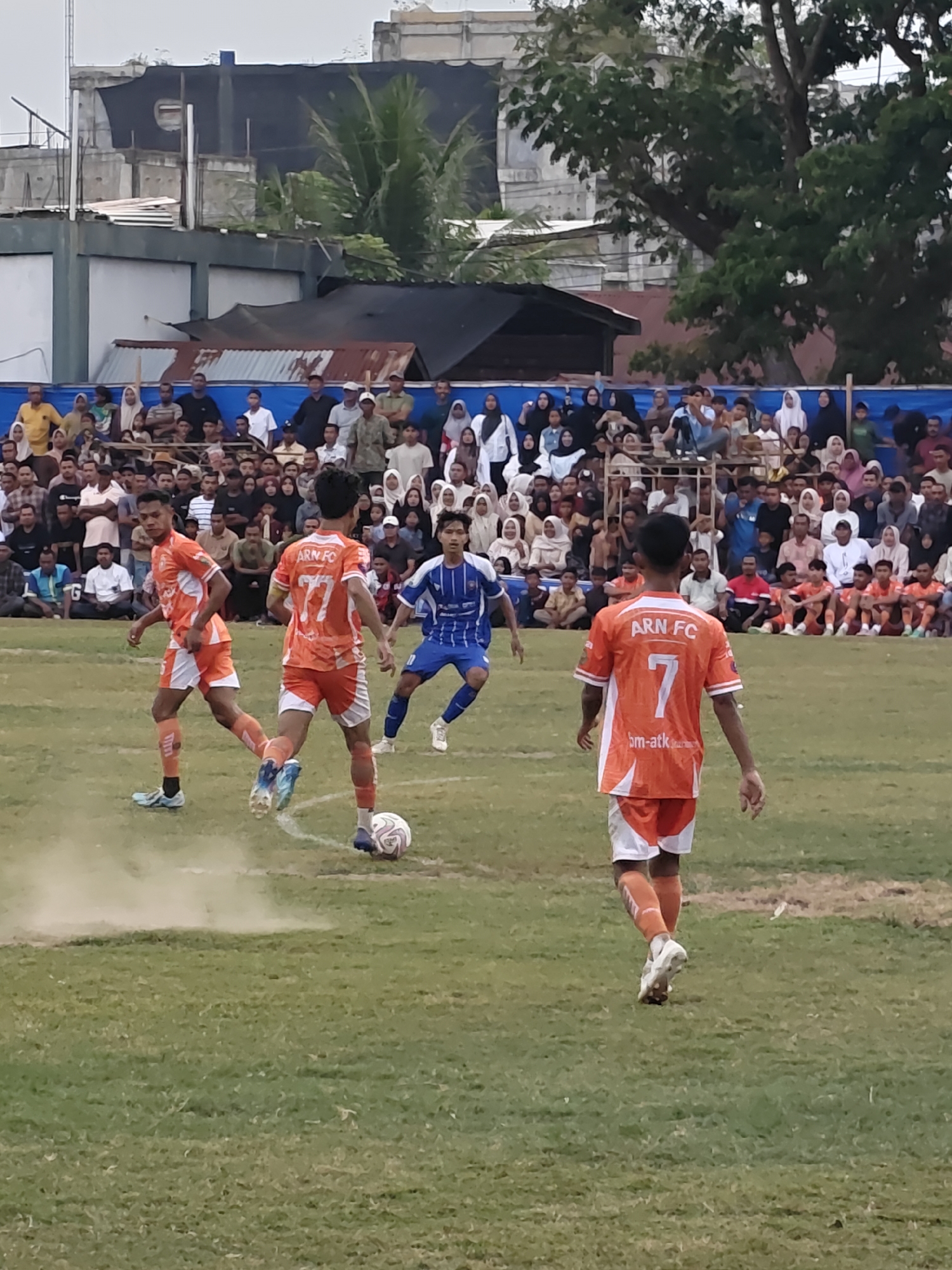This is a medium shot of a soccer game taking place outdoors on a grass field. The game appears to be in a village setting, possibly in Desa Meunasah Blang, Indonesia, given the style of buildings in the background which includes some with corrugated metal roofs and concrete structures. A crowd of spectators is visible behind a low barrier on the right side of the frame.
In the foreground, three male soccer players are in action. Two players are wearing orange jerseys with white accents and white shorts. One of these players, in the left half of the frame, is facing away from the camera, seemingly dribbling the ball. The other orange-jerseyed player, in the center, is also facing mostly away from the camera, with the number "77" visible on his back and "ARN FC" written above it. He is also wearing orange socks. To the right, a third player in a blue and white striped jersey with blue shorts and white socks is facing the orange-jerseyed player with the number "77". A soccer ball is on the ground between the two orange-jerseyed players.
The field is covered in green grass, with some lighter patches and what appears to be a dirt path or kicked-up dust in the immediate vicinity of the players. The background features more spectators, trees, and buildings, suggesting a relatively populated area. The sky is overcast.