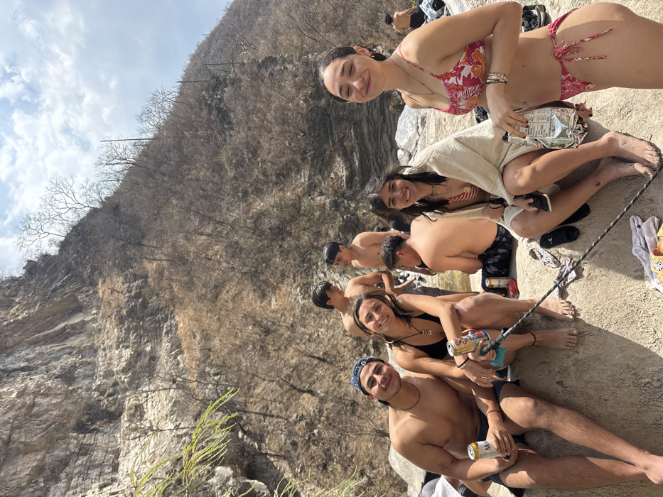 The image captures a group of at least eight young adults in an outdoor, rocky environment in what is identified as San Miguel Atlapulco, Mexico. The foreground features four individuals seated on a light-colored rock surface. From left to right: a shirtless male with dark shorts and a backward dark baseball cap holds a can of Modelo Especial beer, smiling at the camera; beside him, a female in a black bikini top and dark shorts holds a can of Twisted Tea, also smiling at the camera, with a dark pole extending from her vicinity; next, a female in a striped bikini top is partially covered by a light-colored towel and holds a dark object, smiling at the camera; and on the far right, a female in a pink and white floral bikini holds a silver foil packet, with her eyes partially closed and a slight smile. In the mid-ground, three additional shirtless males are visible in or near a shallow body of water, and another male is seated, facing away from the camera. The background consists of a large, steep, light-colored rock face on the left and a dry, barren hillside with sparse, leafless trees and shrubs on the right, under a partly cloudy sky. Scattered clothing items and footwear are visible on the rocks.