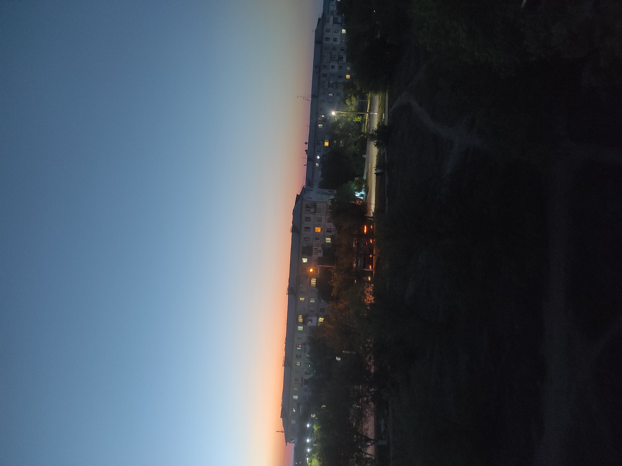 The image captures a twilight scene in the city of Shakhtinsk, Kazakhstan. In the background, a long, multi-story apartment building is visible, with lights on in many of its windows, suggesting it is evening. The sky transitions from a deep blue at the top to a soft orange and pink hue near the horizon, indicative of sunset or twilight. In the foreground, trees and foliage are silhouetted against the fading light, with a dirt path winding through the vegetation. A streetlamp is illuminated, casting a warm glow on the area. Several figures are faintly visible walking on the path in the middle ground. The overall atmosphere is calm and serene, depicting a quiet urban evening.