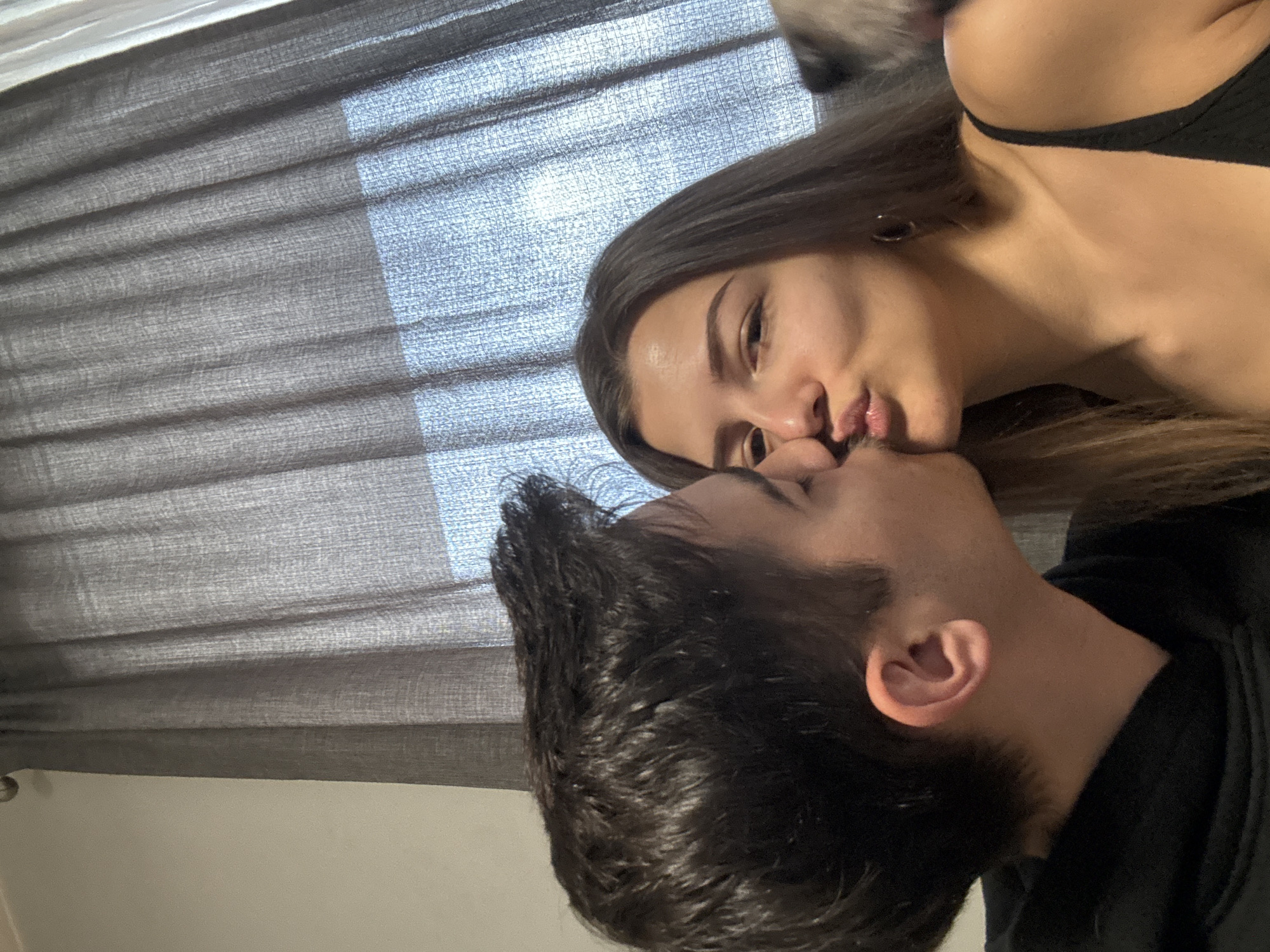 A close-up, high-angle shot shows a young man and woman kissing. The woman, with long brown hair, is wearing a black top. The man has short, dark hair and a beard and is wearing a black hoodie. A gray window blind is visible in the background. The location is not identifiable as being in Sofia, Bulgaria.