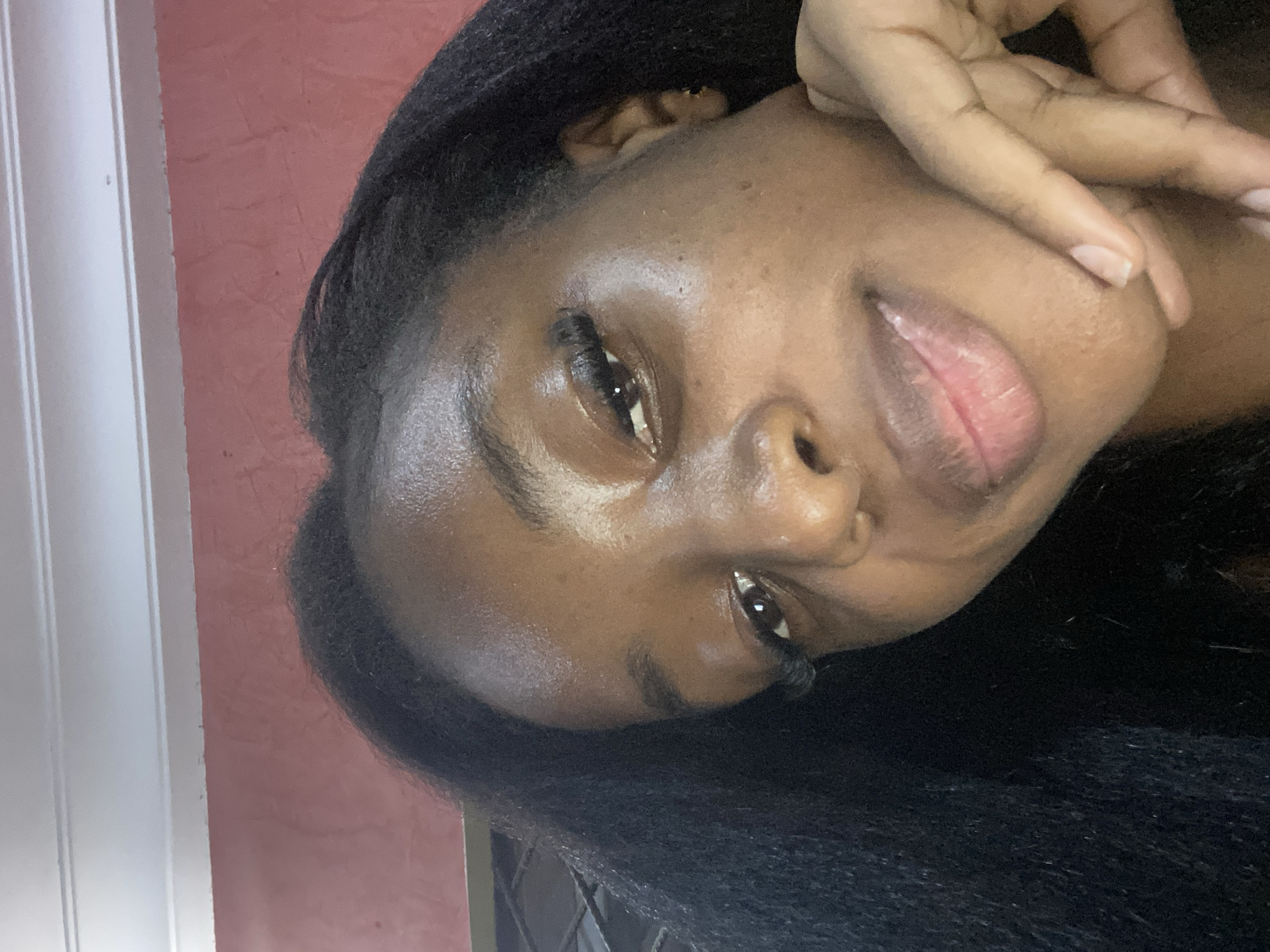 This is a close-up, selfie-style portrait of a young Black woman. She has dark skin, long black hair, and is wearing false eyelashes. Her right hand is resting near her chin, with her fingers gently touching her jawline. The background appears to be an indoor setting with a pink wall and a white ceiling with crown molding. There is no clear indication of the specific location within Jos, Nigeria.