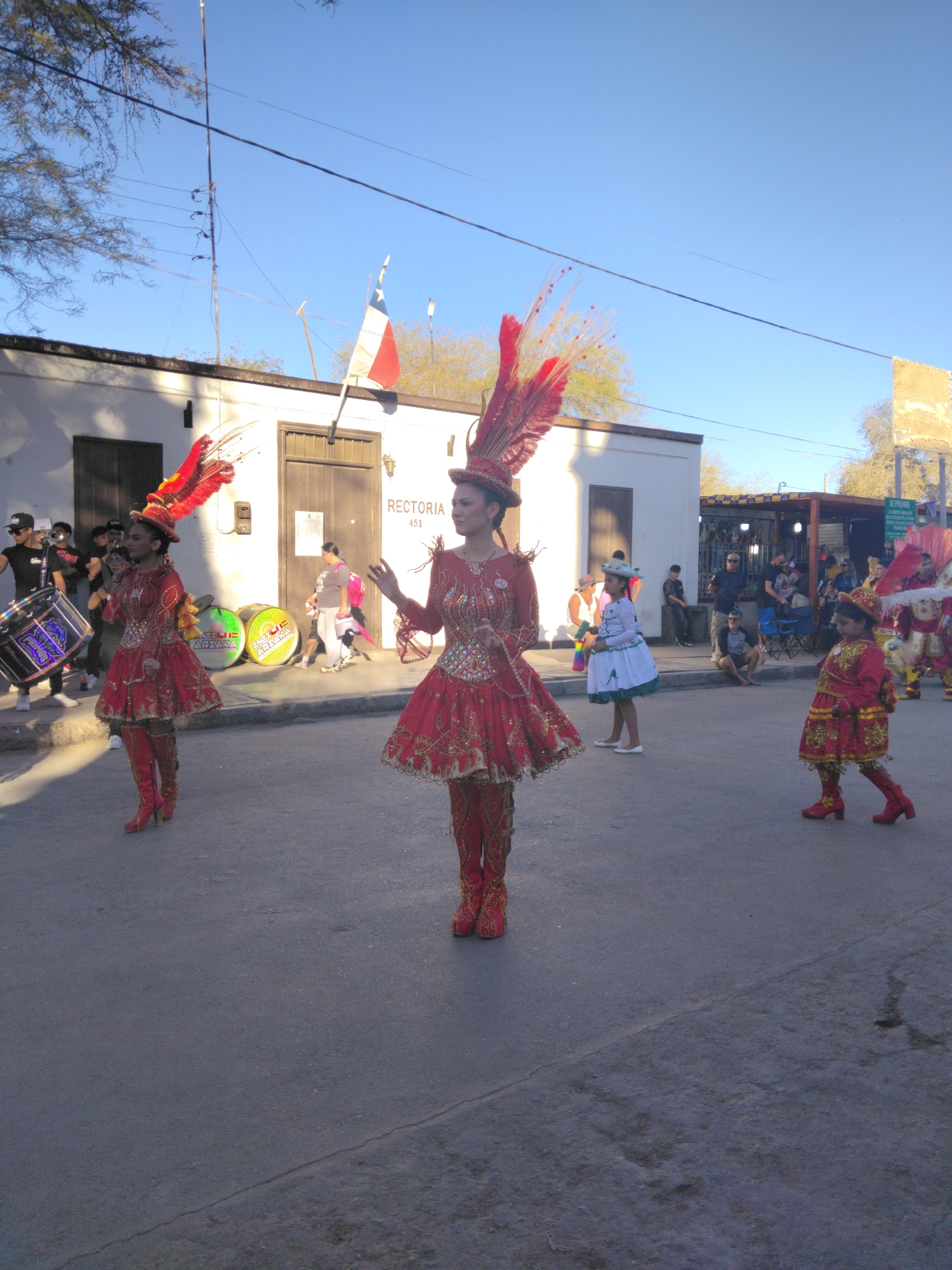 The image depicts a parade in Pozo Almonte, Chile, with participants in elaborate red and gold costumes. In the foreground, three individuals are dressed as performers, two women in matching ornate red dresses with feathered headdresses and tall red boots, and a child in a similar, smaller red costume. The woman on the far left is holding a large drum. Behind them, other participants and spectators are visible. The setting appears to be a street in front of a building with "RECTORIA 451" signage, possibly a municipal building or educational institution, under a clear blue sky. The architecture and arid vegetation suggest a location in northern Chile.