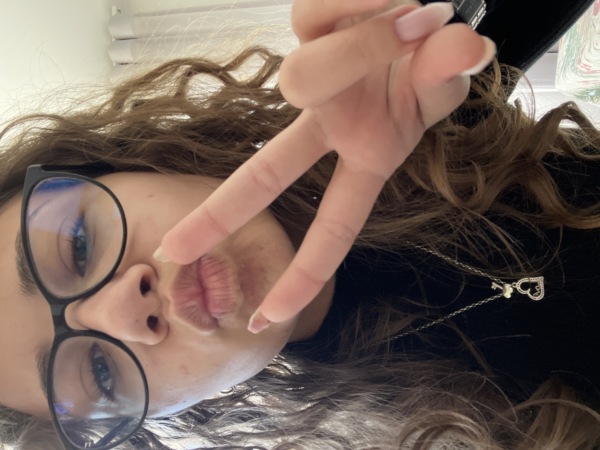 A young woman is posing for a selfie. She is wearing black-rimmed glasses and has light brown, curly hair. She is making a kissing face and holding up a peace sign with her right hand. A silver necklace with a heart-shaped pendant and a key is visible. The background is blurry and indistinct, but appears to be an indoor setting with a white surface. The location of the photo is not discernible from the image. The city of Karavelovo, Bulgaria cannot be confirmed.