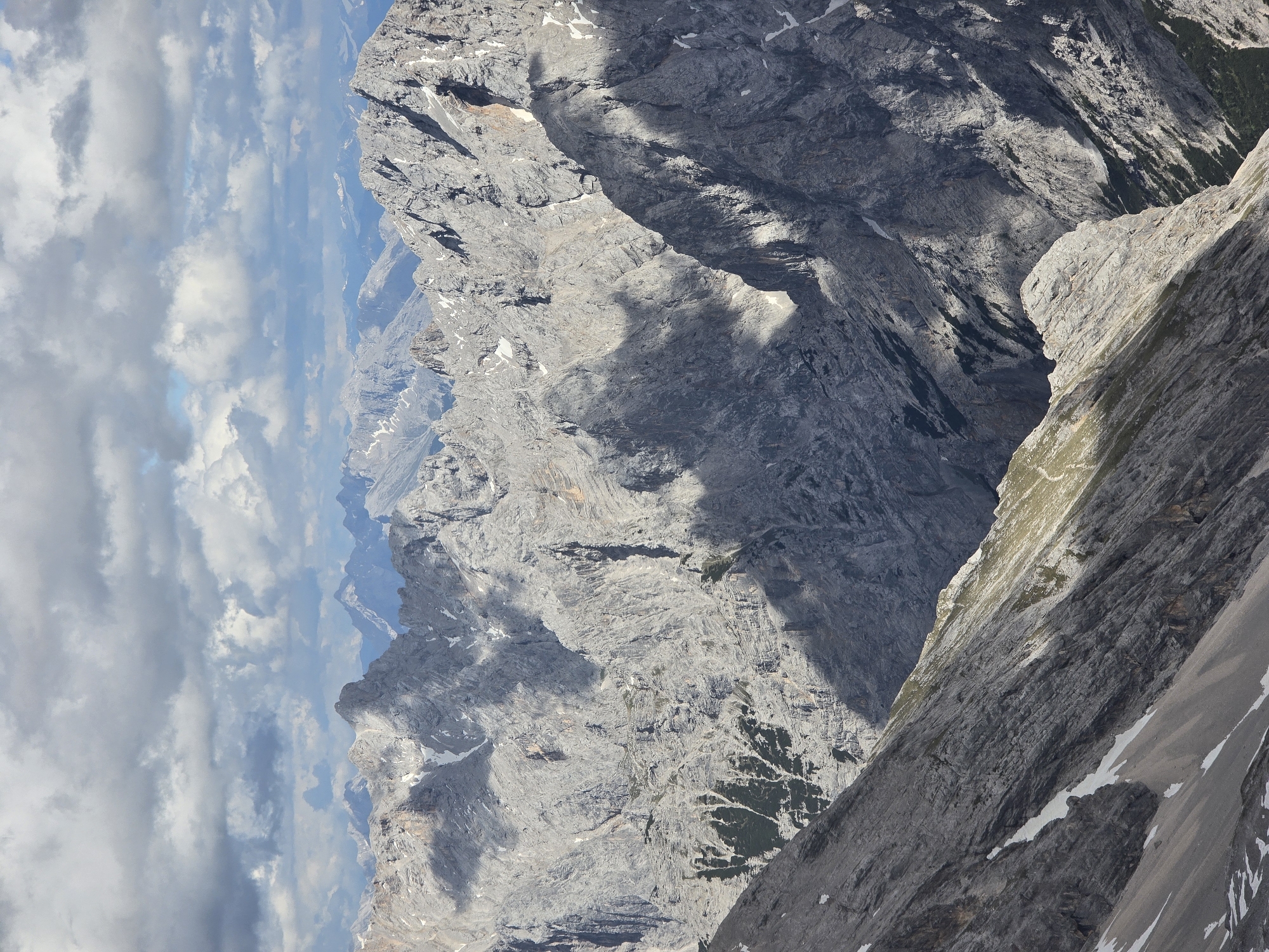 The image is a high-angle, medium shot of a rugged mountain range under a cloudy sky. The mountains are predominantly grey and rocky, with patches of green vegetation in some shaded valleys. The terrain is steep and uneven, characterized by sharp peaks, deep ravines, and scree slopes. Sunlight breaks through the clouds, casting dramatic shadows across the mountainside, highlighting the texture and contours of the rock formations. Small patches of snow are visible on some of the higher slopes. The overall impression is one of a wild and imposing natural landscape. The image appears to be taken in the Bavarian Alps, likely in the vicinity of Garmisch-Partenkirchen, Germany, given the characteristic geological features of the region, such as the jagged limestone peaks and alpine vegetation. There are no people, buildings, or man-made objects visible in the frame.