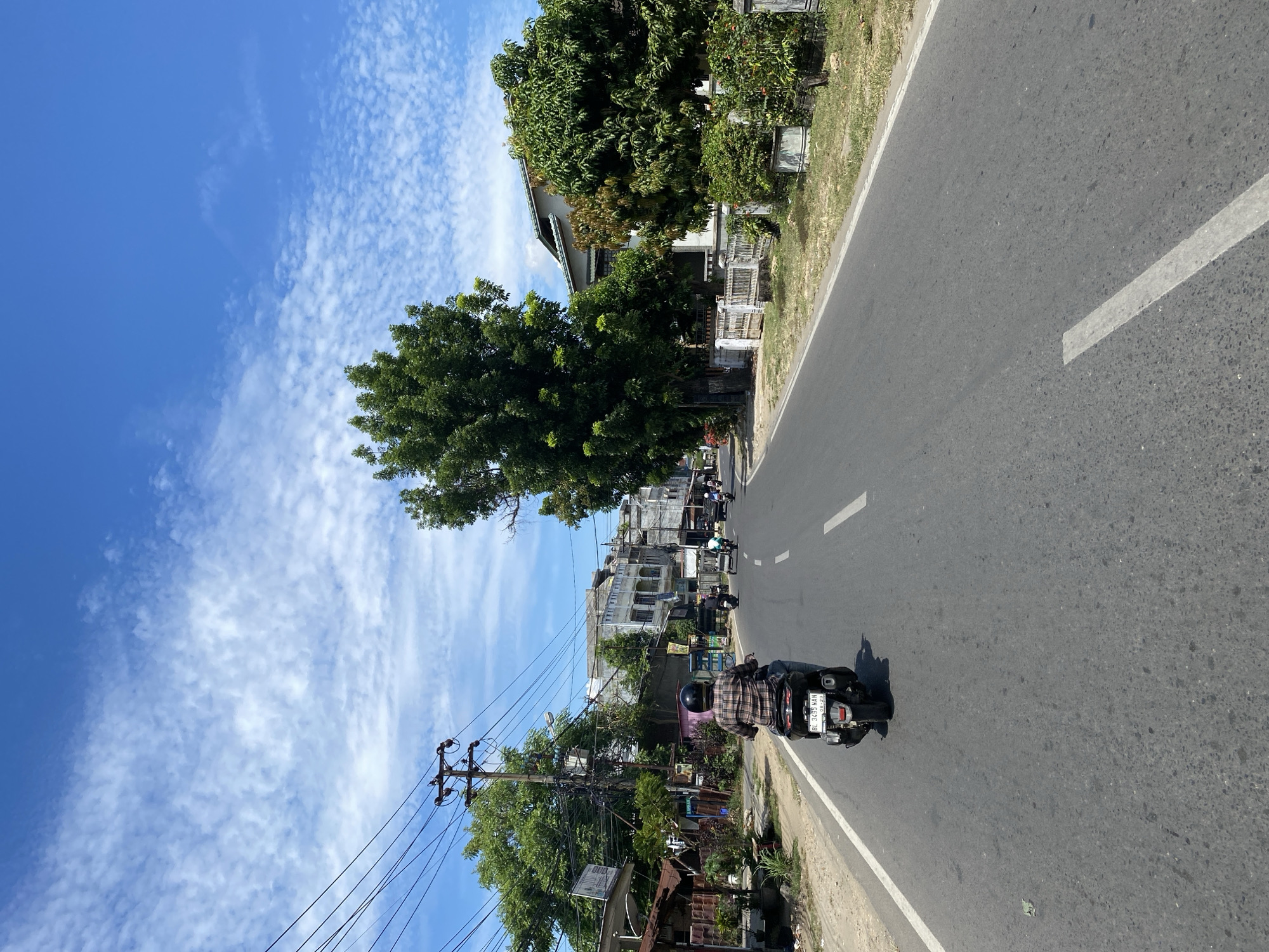 A person is riding a scooter down a paved road in Lhokseumawe, Indonesia, on a sunny day with scattered clouds. The rider, wearing a plaid shirt and helmet, is in the foreground, moving away from the camera. The road has white lane markers. On the left side of the road, there are utility poles with many wires, trees, and buildings. On the right side of the road, there are houses, fences, and lush green trees. Other vehicles, including another scooter and a three-wheeled vehicle, are visible further down the road. The sky is a vibrant blue with white, wispy clouds.