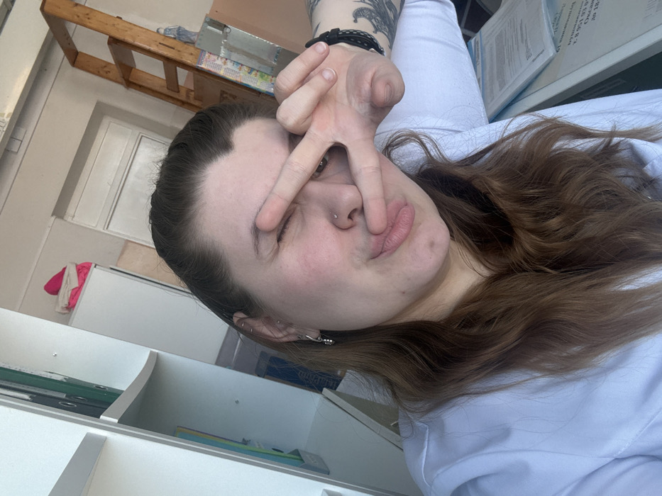 A person with long brown hair poses for a selfie while winking and making a peace sign over one eye. They have a nose piercing and wear multiple earrings. The background includes a white wall and a light-colored shelf with colorful stationery. A wooden structure and white cabinets are visible further behind. The person is wearing a light-colored top.