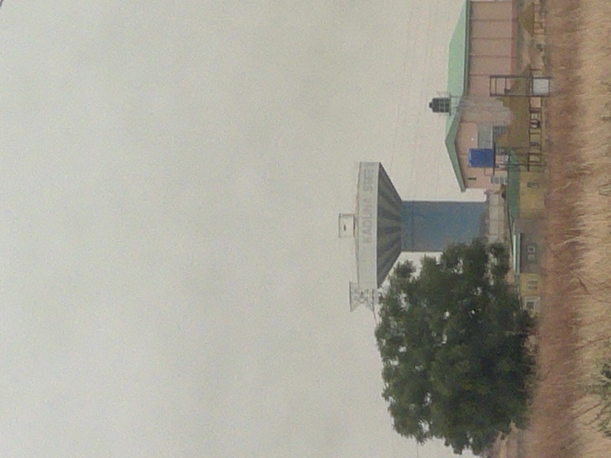 The image shows an industrial area in Afaka, Nigeria. Dominating the scene is a tall, blue and gray structure with "KADUNA STEEL" vertically written on it. A green tree is located near the structure. To the right, there are a few buildings with green roofs. The ground is covered in dry grass. The sky is overcast, with gray haze.