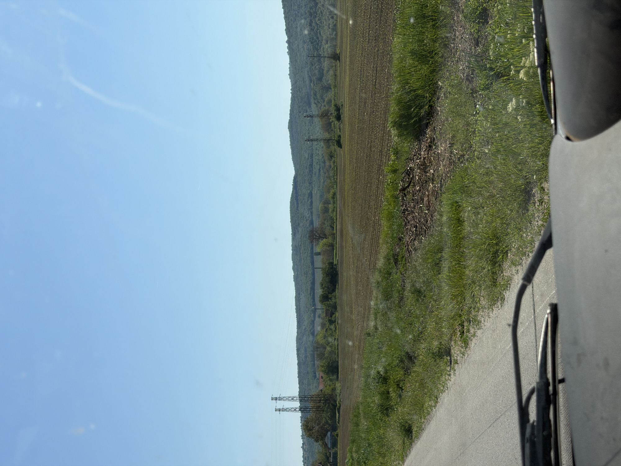 The image shows a rural landscape, seemingly taken from a moving vehicle. A clear blue sky dominates the left side of the frame. To the right of the sky, there is a field of freshly tilled soil, bordered by a strip of green grass and vegetation. A tree-covered hill or mountain range runs parallel to the field. Power lines with metal towers extend from the bottom of the frame towards the middle distance, spanning the field. A road runs along the bottom edge of the image, curving towards the right side. Part of a vehicle's side mirror and frame can be seen on the extreme right, partially obscuring the view. Based on the information in the query, the location is Ioglav, Bulgaria, although there are no specific features definitively linking the scene to that precise place. The scene lacks any immediately identifiable people or interactions.
