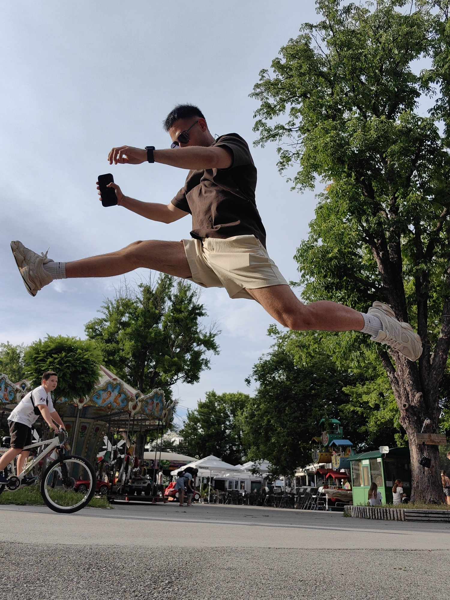 An individual, dressed in a brown t-shirt and light-colored shorts, is captured mid-air in a dynamic split-jump pose. They are holding a black smartphone in their right hand and appear to be wearing sunglasses and a smartwatch. The individual is positioned against a backdrop of a clear sky and lush green trees. In the lower portion of the frame, a young person is riding a bicycle, and a carousel with colorful decorations is visible, suggesting a park or amusement area. The ground in the foreground is paved with gravel. The scene is likely set in the city of Shumen, Bulgaria, given the context of the carousel and park-like surroundings, which are common features in public spaces within the city.