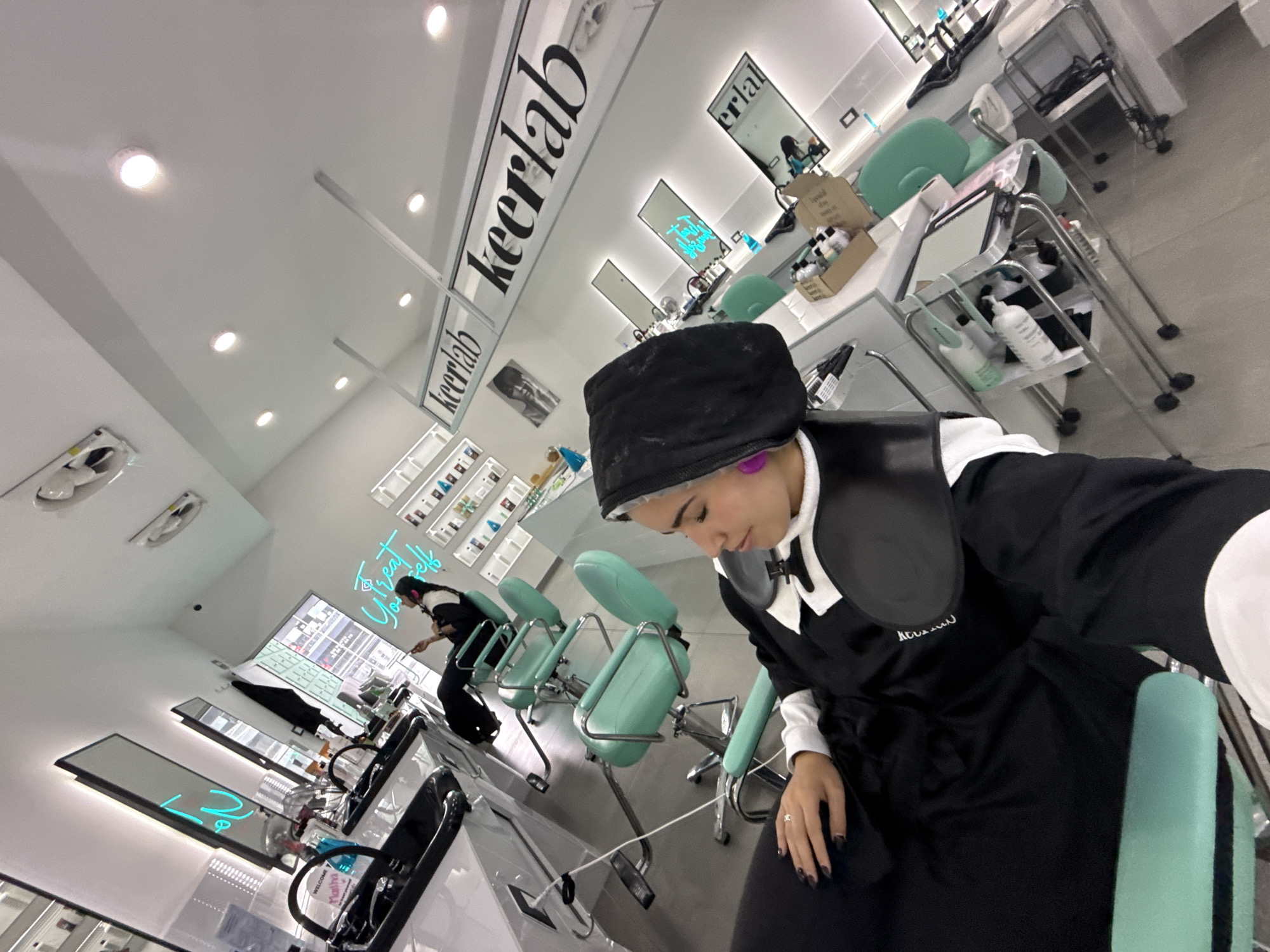 Here is a description of the image:
The photo captures the interior of a hair salon, likely located in San Bernardino Tlaxcalancingo, Mexico. The dominant feature is a woman in the foreground seated in a salon chair, covered in a black gown. She is wearing a black head covering. In the background, another woman is also seated in a salon chair, presumably receiving hair treatment. The salon itself is modern and well-lit with light green chairs and metallic equipment. "keerlab" is written in black on the ceiling, suggesting the name of the salon. The image is taken from an oblique angle.