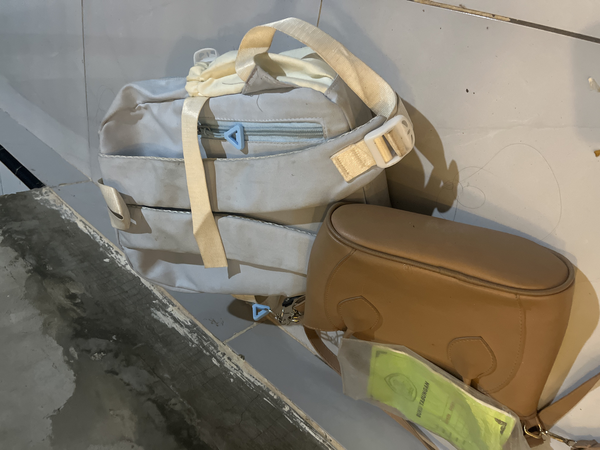 Two bags, a light grey backpack and a brown shoulder bag, are on a tiled floor in an indoor environment. The backpack has beige straps and a zipper with a blue triangular pull. The shoulder bag is made of leather and has a strap attached. A clear plastic sleeve containing a green booklet with Indonesian text is attached to the shoulder bag. The background consists of a grey, rough-textured wall and a white tiled floor. The scene is lit from above, casting shadows. The location is identified as Kebumen, Indonesia.