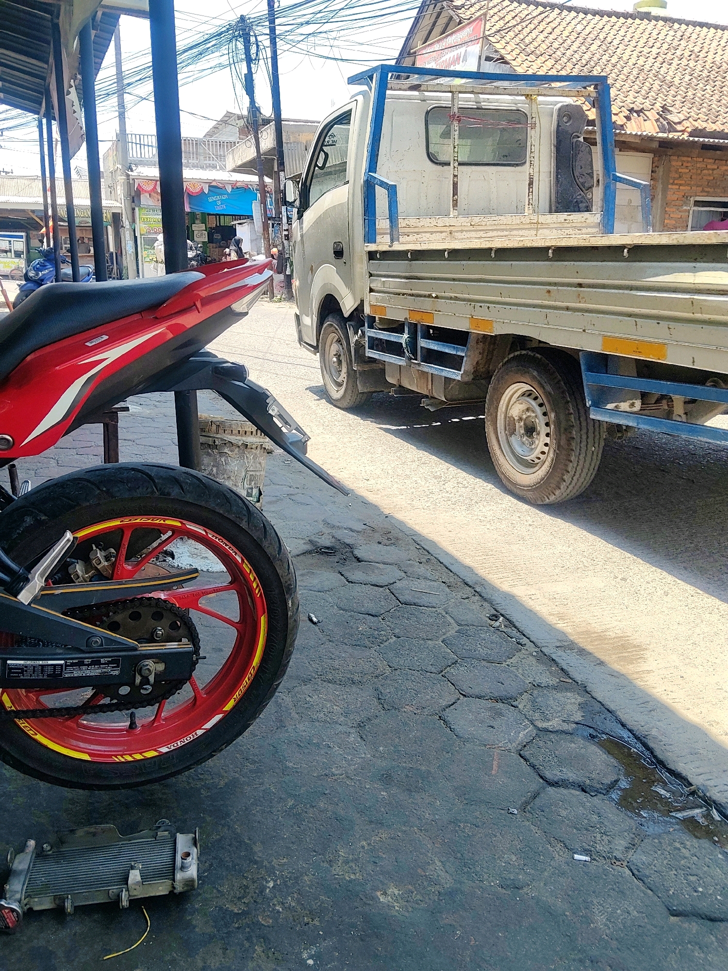 The image depicts a roadside scene in Mekarmukti, Indonesia, under a bright, sunny sky. In the foreground, a red and black motorcycle is parked, its rear wheel prominently displayed with a red rim and yellow decals. To the right of the motorcycle, a light-colored flatbed truck is parked on the street. The truck has a blue metal frame over its bed and is positioned next to a sidewalk constructed of hexagonal paving stones. Beyond the truck, buildings with tiled roofs and various signage are visible, suggesting a commercial or residential area. Overhead, a dense network of electrical wires crisscrosses the sky. In the lower left of the frame, a detached motorcycle radiator lies on the ground. The ground surface appears to be a mixture of asphalt and paving stones, with some water and debris present. There are no people clearly visible in the frame.
