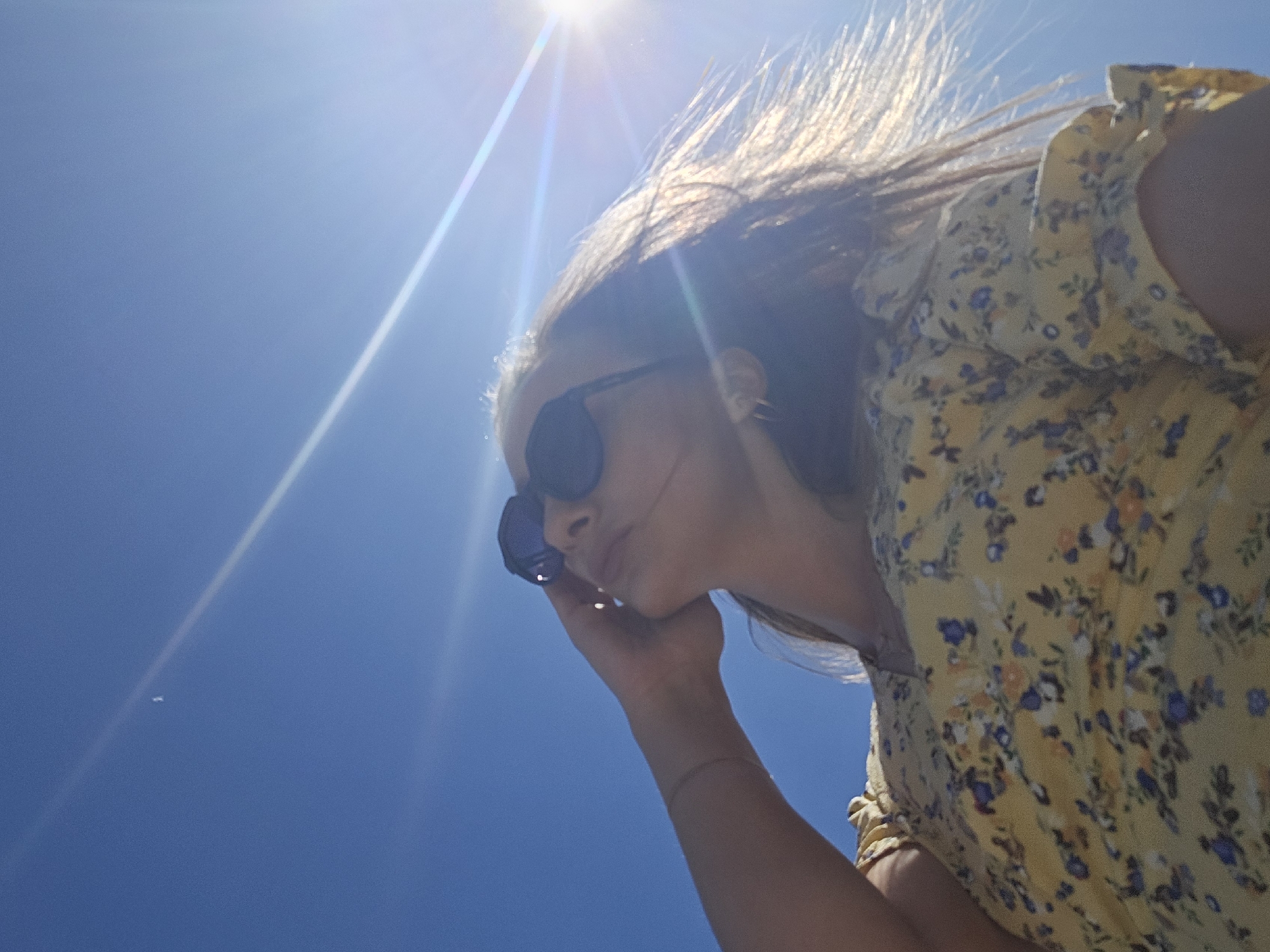 A woman wearing sunglasses and a yellow floral dress is positioned against a bright blue sky with the sun shining overhead. The location cannot be determined based on the image. The woman's hand is near her face, and her hair is blowing in the wind.