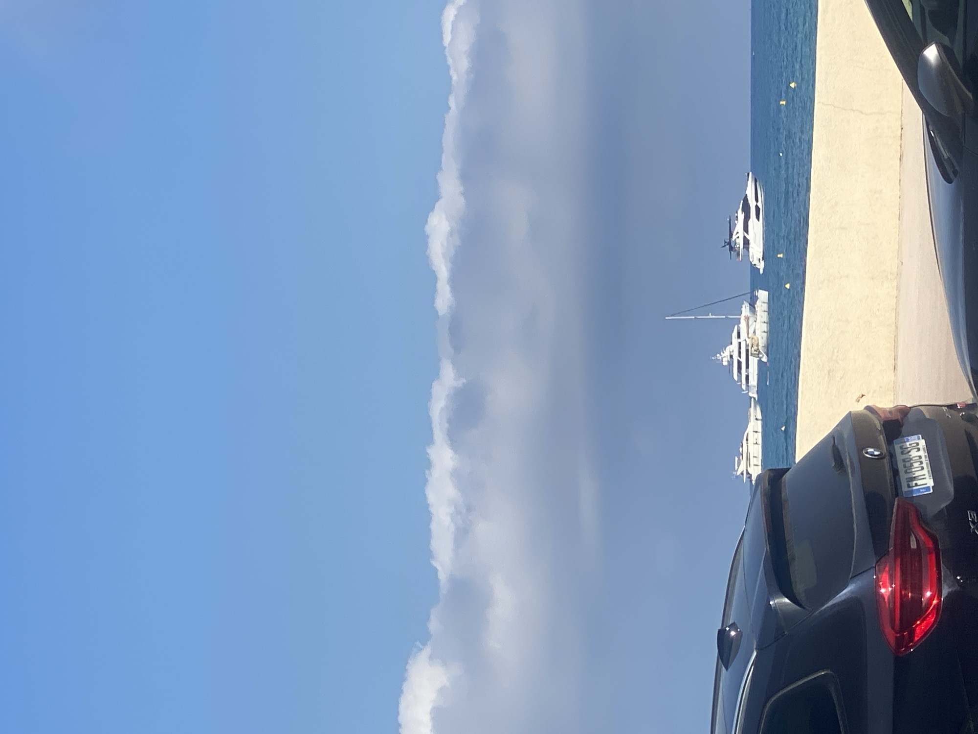 In Antibes, France, a black BMW SUV is parked in the foreground, facing away from the viewer. Its taillights are visible, along with the license plate "FM-058-SG". Beyond the SUV is a sandy beach, and further out, the Mediterranean Sea. Several yachts and a sailboat are anchored on the water under a bright blue sky with a line of white clouds. Another dark-colored car is partially visible in the lower right corner of the image. The scene depicts a sunny day by the coast.