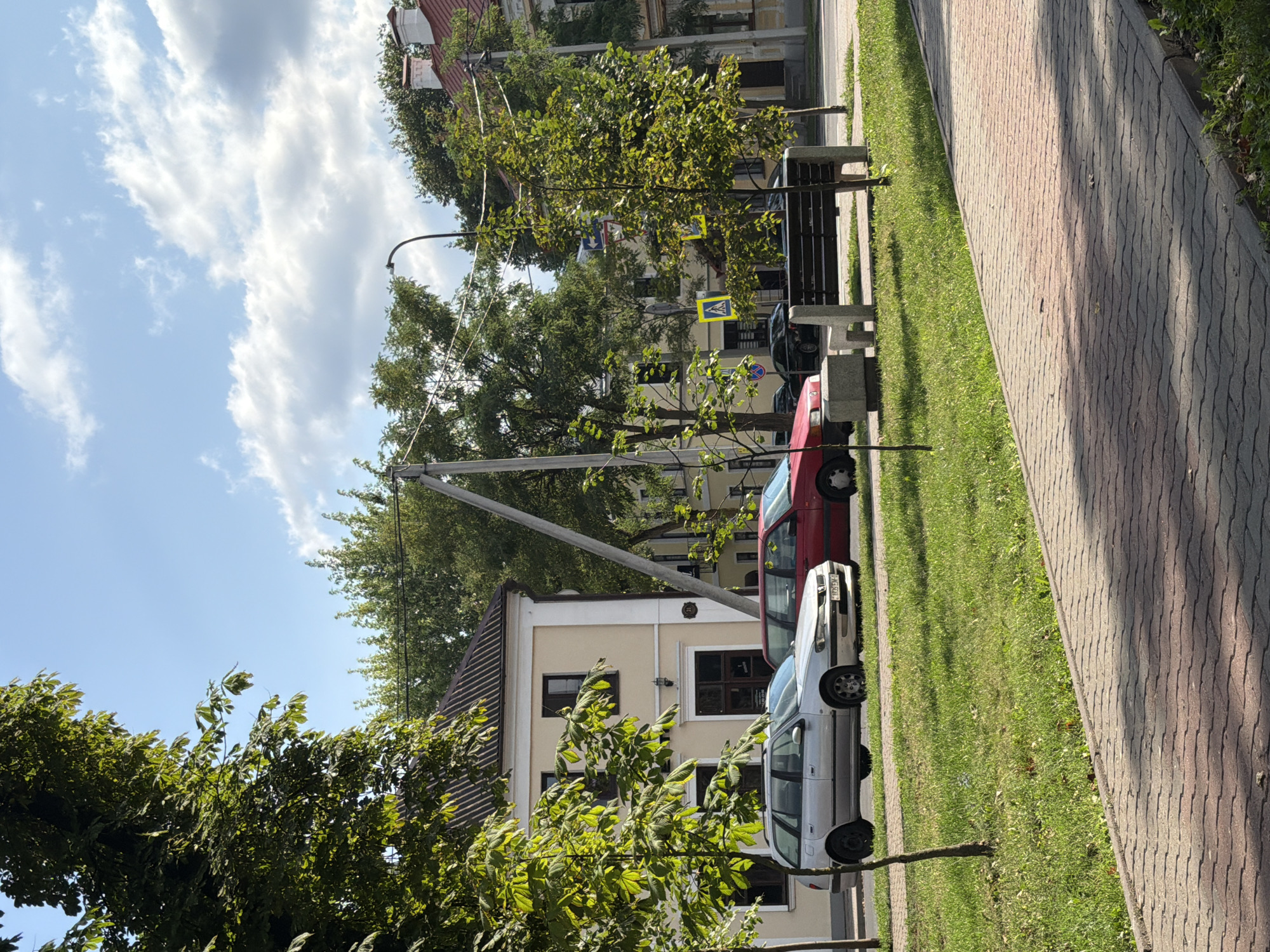 The image depicts a street scene in Brest, Belarus, on a sunny day. In the foreground, a paved walkway runs diagonally across the frame, bordered by a grassy area. A young tree stands in the grass to the left. To the right, a park bench with stone supports and a wooden seat sits on the grass, with a small tree behind it. In the middle ground, a silver station wagon and a red sedan are parked on the side of the road. Behind the cars, a light-colored building with a dark tiled roof is visible, along with other buildings and mature trees lining the street. Power lines and a street lamp are also present in the scene. The sky is blue with scattered white clouds.