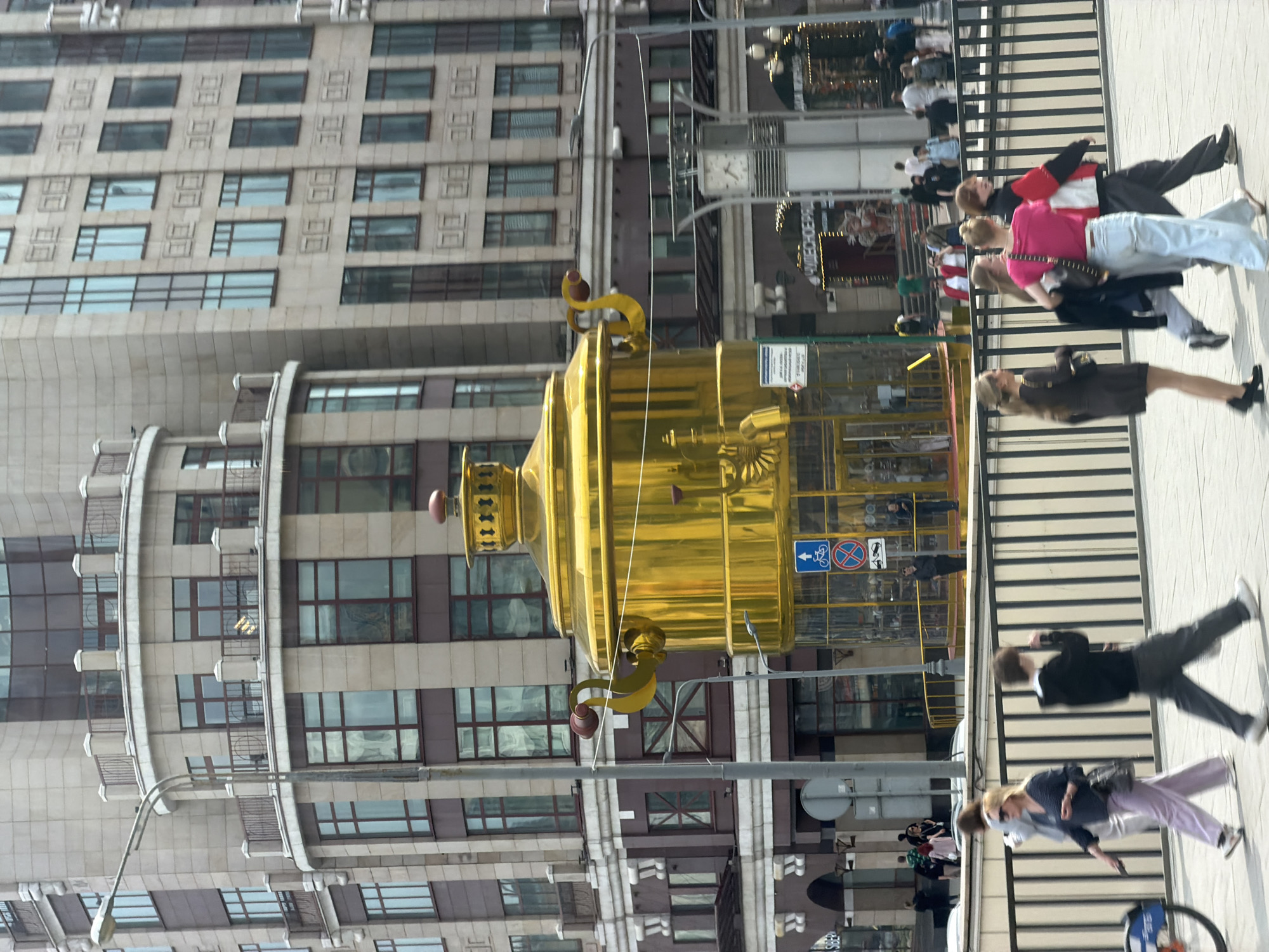 The image shows a large golden samovar structure in the center of Moscow, Russia. The surrounding environment includes a modern building with many windows, a clock tower, a fence, a street crossing, and pedestrians walking. The scene appears to be in a public area, likely a square or street where people are passing by.