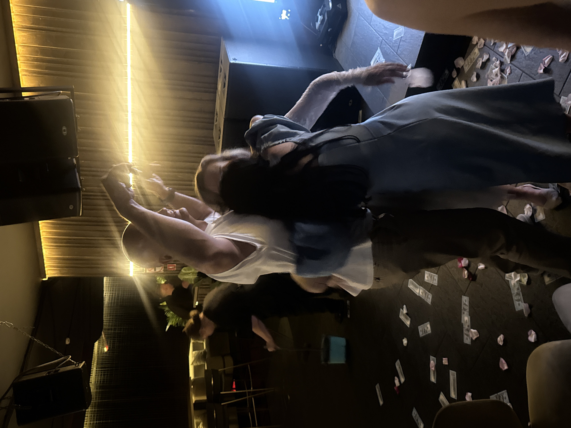 This image depicts an indoor scene, possibly a nightclub or event venue, with a dark ambiance illuminated by golden spotlights and ambient lighting. In the foreground, two individuals are interacting. One, wearing a white t-shirt and dark pants, has their arms raised, holding something unseen. Another individual, dressed in a light blue dress and white gloves, stands close by, facing away from the camera, with their arm extended. The floor is littered with what appears to be money and rose petals. In the background, a person in a black uniform is using a broom and dustpan to clean the floor, suggesting a post-event cleanup. The environment includes speakers suspended from the ceiling and a stage or raised platform. The presence of money and celebratory elements on the floor could indicate a recent party or event. The image does not contain any elements that definitively place the scene in Krasnodar, Russia.