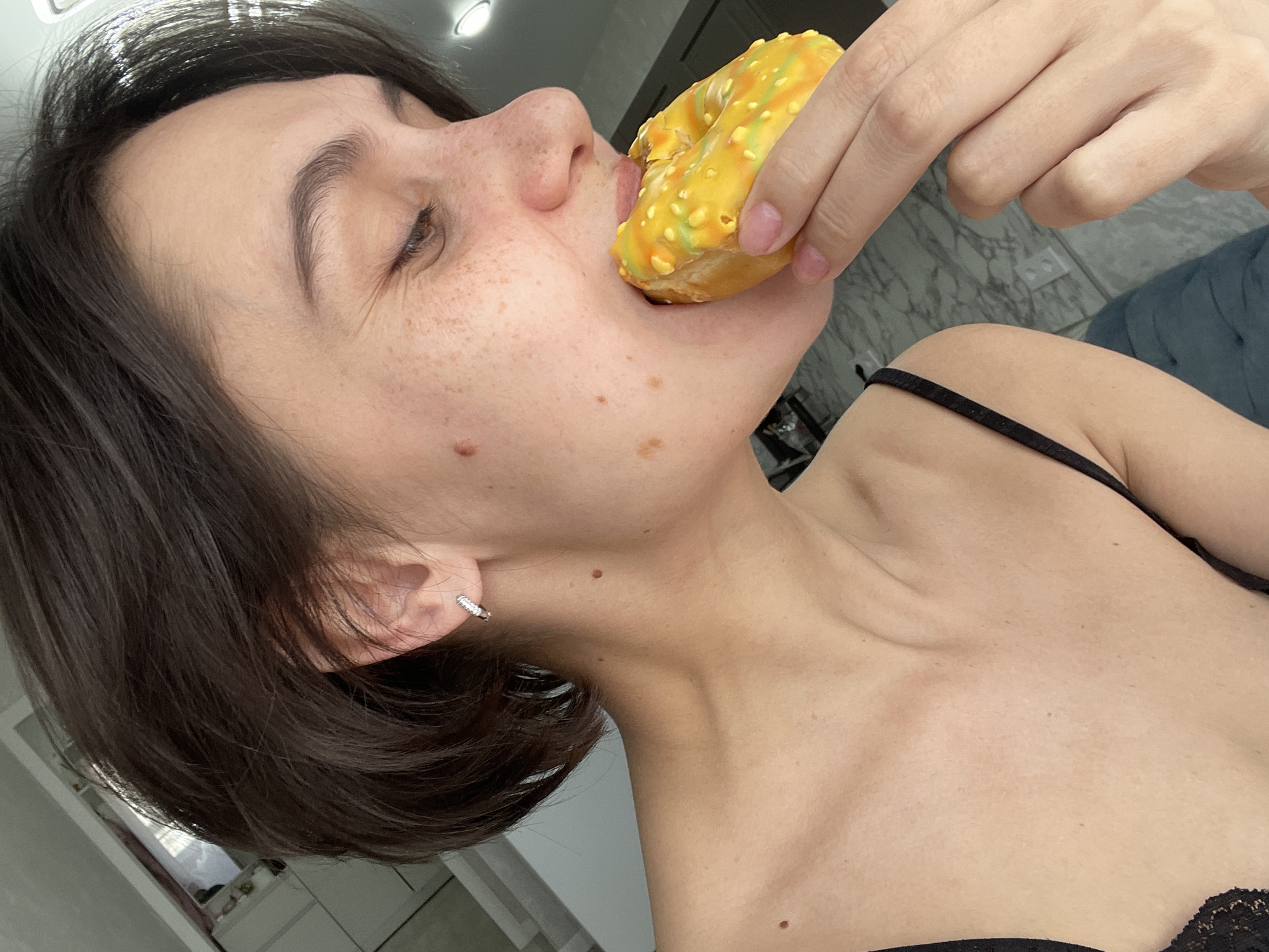 A woman with short brown hair is holding a yellow glazed doughnut to her mouth, biting into it. She has freckles and a mole on her face and a diamond earring in her left ear. She is wearing a black camisole top. The background shows a modern bathroom with marble tile and white cabinets. The location is not discernible as Izhevsk, Russia based on the visual information provided.