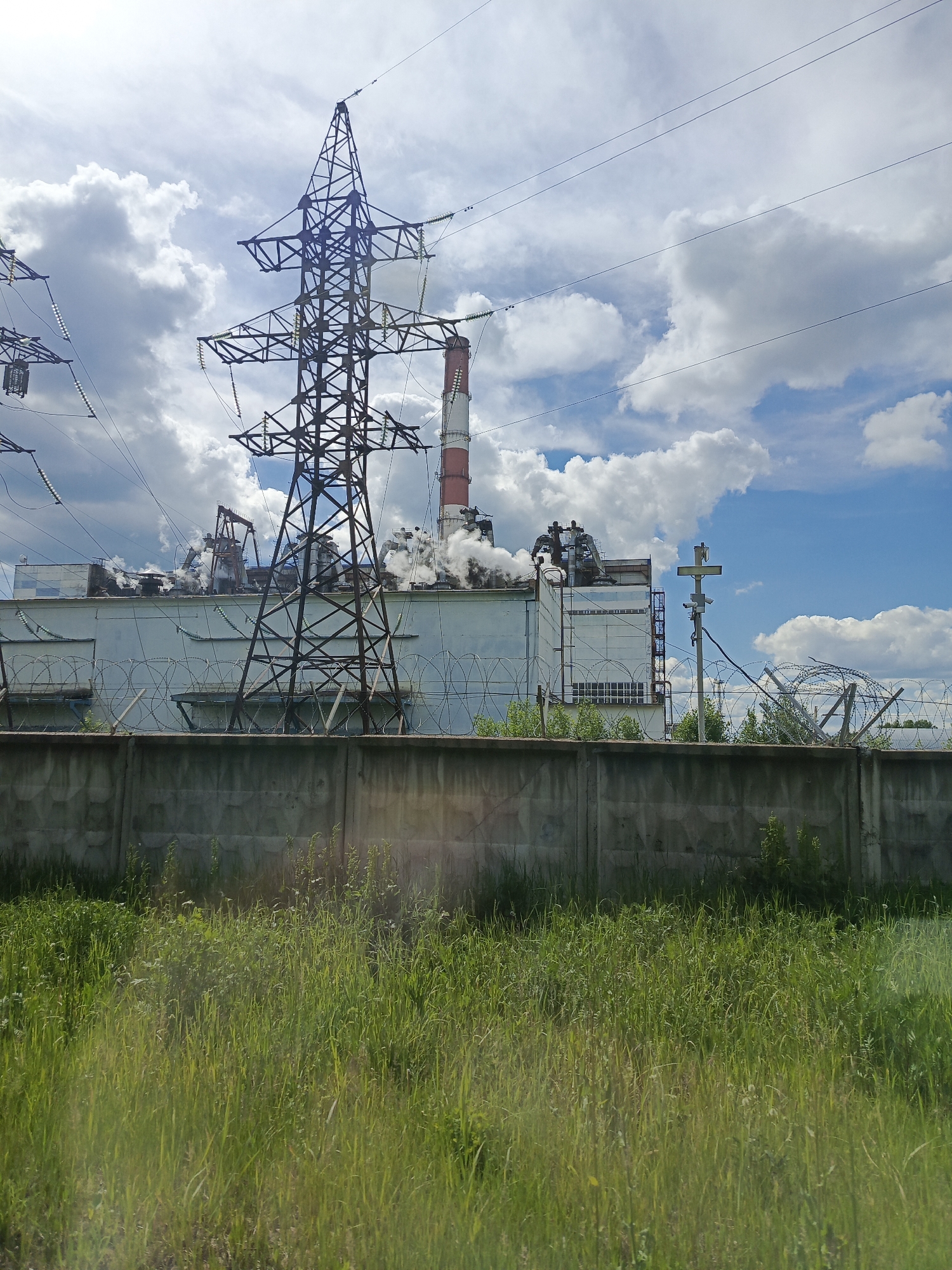The media file displays an industrial landscape under a partially cloudy, bright blue sky. The central subject is a large industrial facility, likely a power plant or factory, featuring a light-colored building with a long, red and white striped smokestack. White smoke or steam is actively being emitted from multiple vents and structures on the roof of the building, indicating ongoing operation.
In the foreground, a field of tall, green grass occupies the lower third of the image. Behind the grass is a sturdy concrete fence, topped with menacing rolls of barbed or razor wire, suggesting a secured perimeter. Several large, latticed metal electricity pylons (transmission towers) with insulators and power lines dominate the upper and mid-ground, conveying the transmission of electricity. One prominent pylon stands directly in front of the factory building. To the far right, behind the fence, a utility pole with what appears to be a camera or sensor system is visible.
The sky above is a mix of fluffy white cumulus clouds and clear blue patches, suggesting fair weather during daytime. No people are visible in the image, and there is no discernible text on any of the structures. The overall impression is one of an active industrial site, possibly a power generation facility, secured from public access.