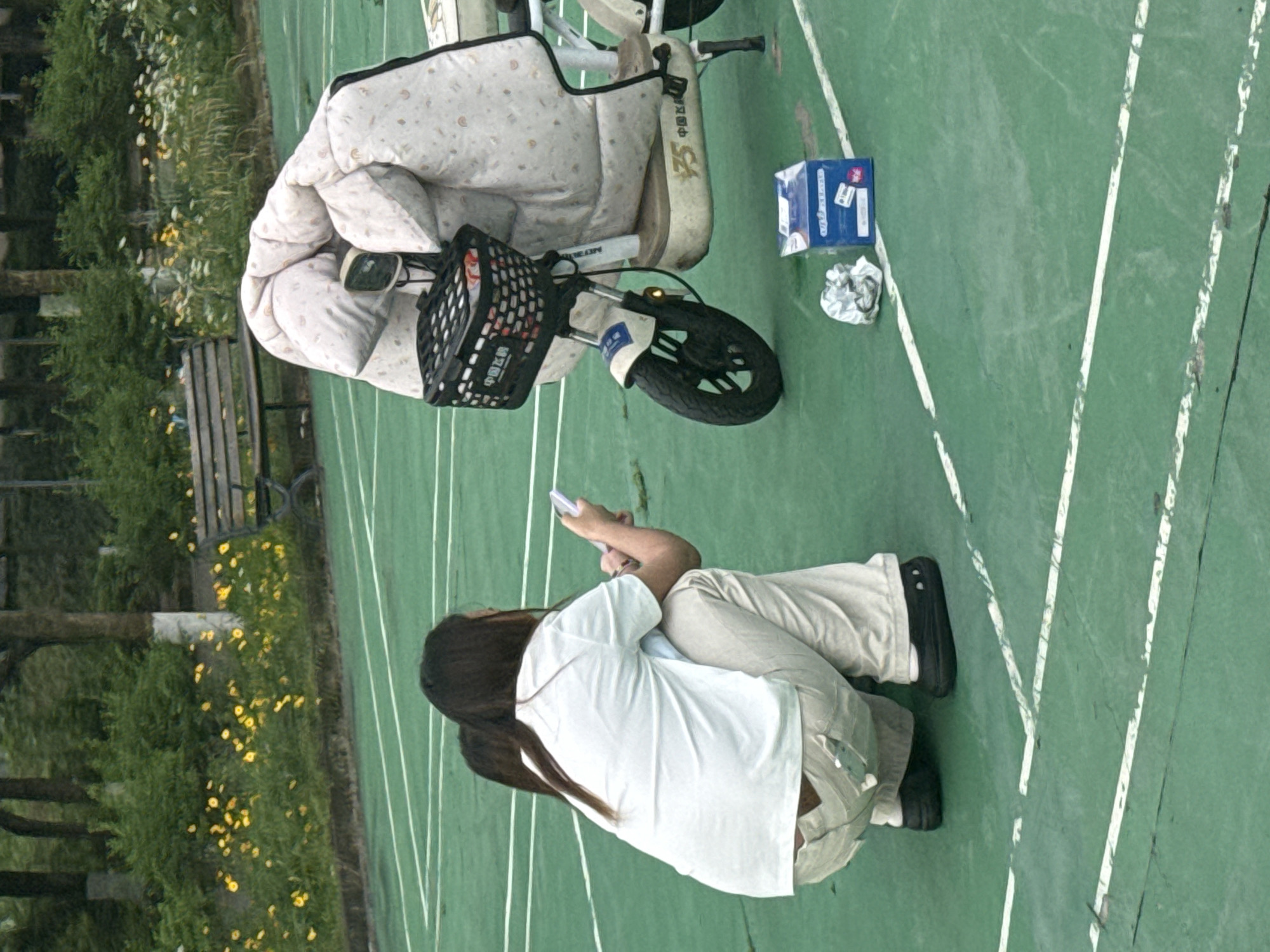 The image shows an outdoor scene in what appears to be Jiangjin, China, based on the Chinese characters on the bike. A woman is squatting on a green surface marked with white lines, possibly a sports court or public area. She is looking at a mobile phone in her hands. Next to her is a small electric bicycle with a padded seat cover. The bike has a front basket and a small box sitting next to it. In the background, there is a bench and trees with yellow flowers. The scene appears to be a public park or recreational area.