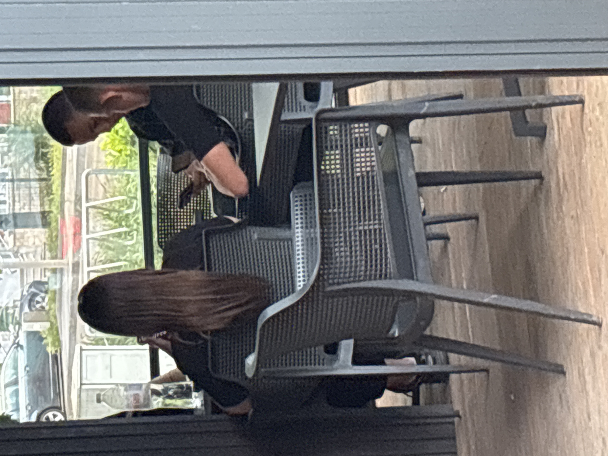 A couple is seated at a metal mesh table and chairs in an outdoor setting, observed from behind what appears to be a window or doorway. The man has short, dark hair and is wearing a black shirt. The woman has long brown hair and is also dressed in black. A water bottle sits on the table. Outside the window/door, a street scene is visible, featuring parked cars. The setting may be located in Plovdiv, Bulgaria.