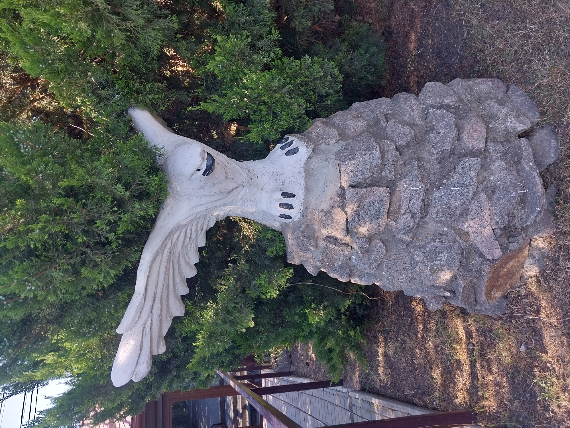 The image shows a stone statue of an eagle perched on a rock pedestal, situated in front of a dense green evergreen hedge. The eagle statue is white with black accents for its eyes and claws, which are depicted as oval shapes. The eagle has its wings spread open, as if in mid-flight or about to take off. The rock pedestal is constructed from irregularly shaped gray stones. To the left of the statue, there is a paved pathway with a dark metal railing. The ground around the statue and pathway is covered in dry grass and dirt. The overall scene appears to be outdoors, likely in a park or garden setting. This is the sculpture of an eagle at the entrance to the Petrykivka Art Museum in Petrykivka, Ukraine.