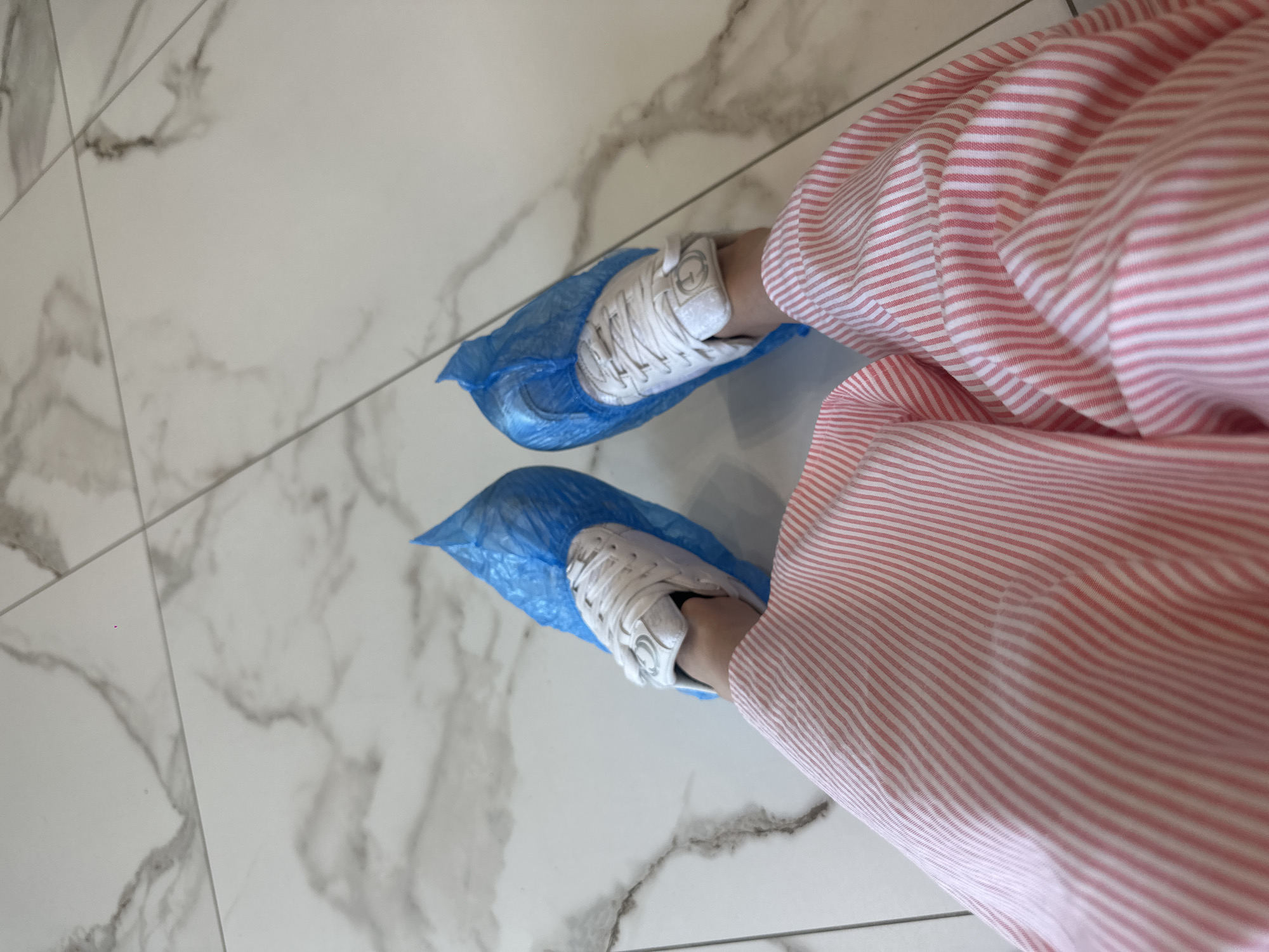 A first-person perspective shot of someone's legs and feet, wearing white sneakers covered in blue shoe covers. The individual is wearing pink and white striped pants. The shoes and pants are resting on a marble-tiled floor. The image appears to be taken indoors, possibly in a medical facility or a place requiring shoe covers.