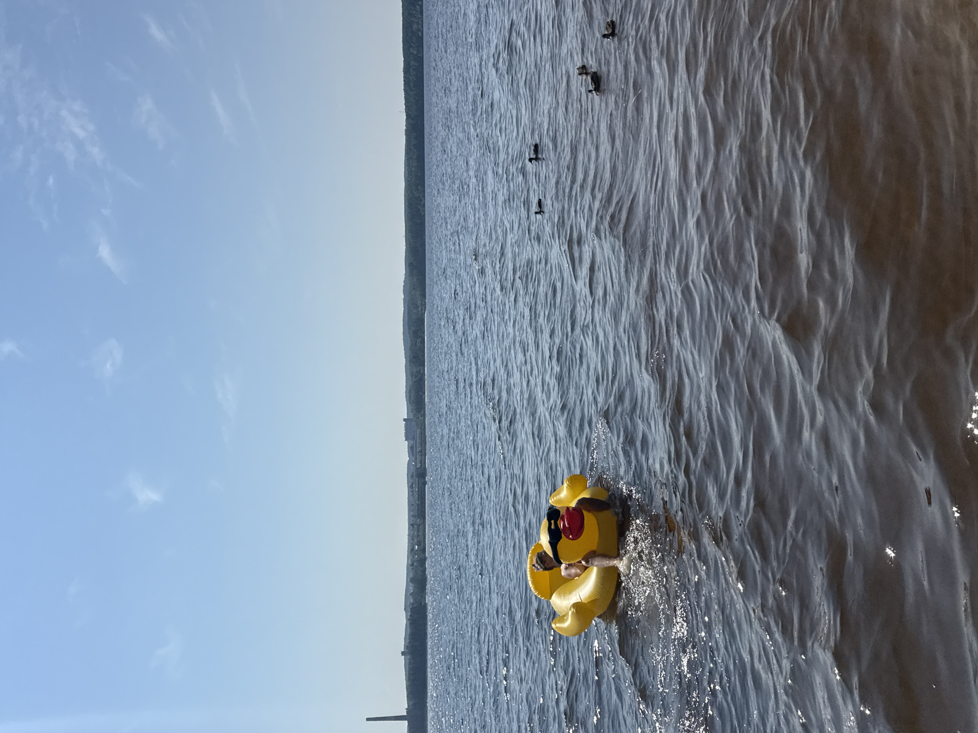A person is floating on a bright yellow inflatable duck in a large body of water. The person is wearing sunglasses and has their legs extended in the water, creating splashes. Several ducks are swimming in the water in the background. The water is choppy, with sunlight reflecting off the surface. In the distance, a shoreline is visible with trees and some buildings, including a tall industrial smokestack on the left. The sky is clear and blue with a few wispy clouds. This scene is located on the Kama River in the city of Chaikovsky, Russia.