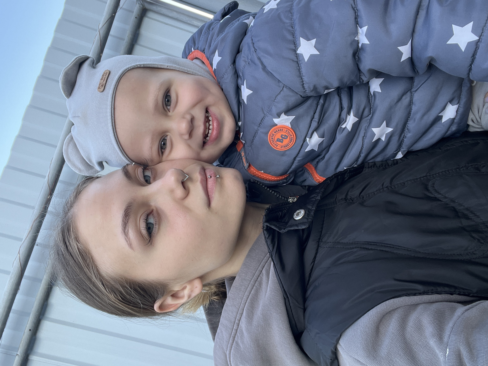 A woman and a child are posing for a selfie. The woman has light skin, light eyes, and light brown hair pulled back from her face. She has a nose ring and a lip piercing. She is wearing a gray hooded sweatshirt and a black puffer vest. The child is lying on top of her. The child has light skin, light eyes, and a few visible teeth. The child is wearing a gray hat with bear ears and a blue puffer jacket with white stars. The background appears to be a metallic corrugated roof or wall. The scene is outdoors, with a hint of blue sky visible in the upper left corner. There is no information in the image to indicate the location is Фоки, Russia.